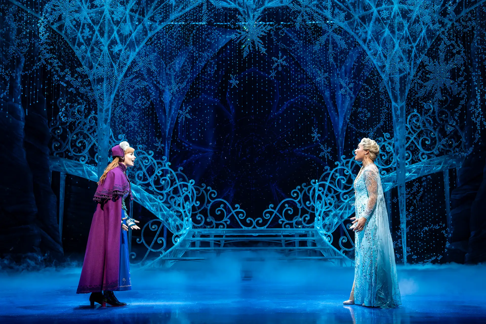 Zwei Frauen in Bühnenkostümen stehen sich auf einer Bühne mit eisigem, blau beleuchtetem Bühnenbild gegenüber.