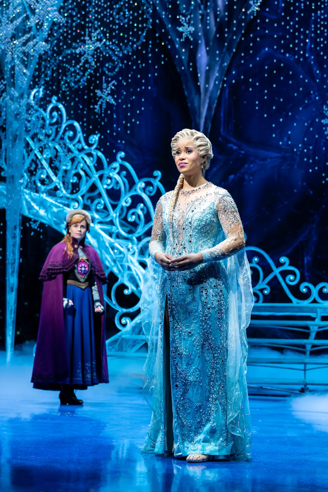 Zwei Schauspielerinnen in aufwändigen Kostümen auf einer Bühne mit eisigem, funkelndem Bühnenbild, eine trägt ein langes, hellblaues Kleid mit transparenten Ärmeln, die andere ein blaues Kleid mit lila Umhang.