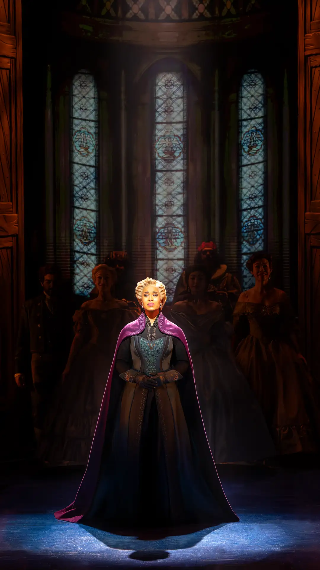 Elsa steht in einem langen Kleid und Umhang vor hohen, verzierten Kirchenfenstern auf einer Bühne im Musical 