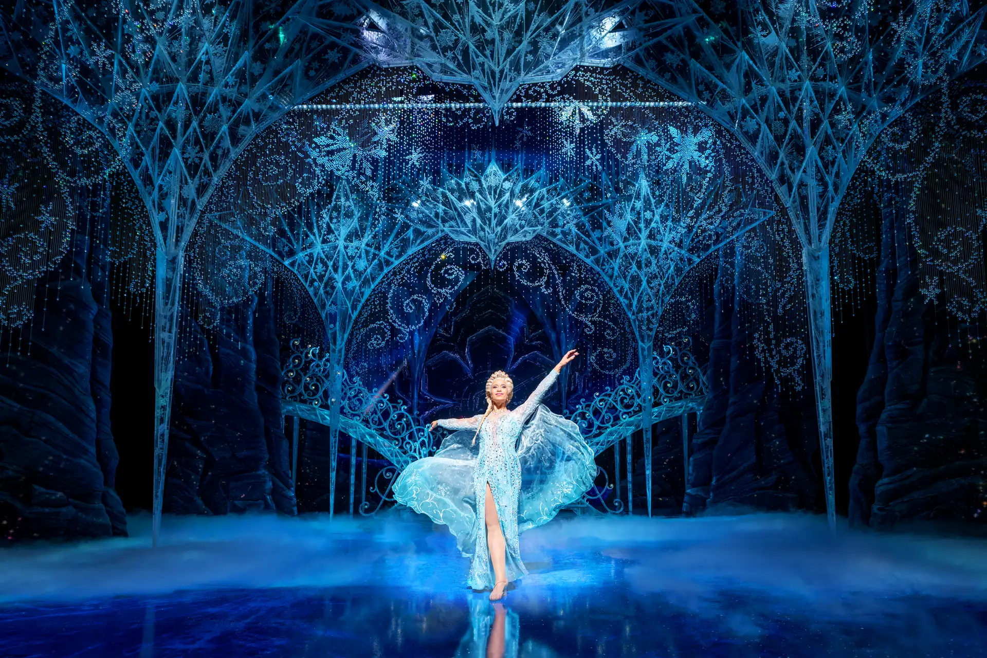 Eine Tänzerin in einem glitzernden, eisblauen Kleid mit ausgestreckten Armen auf einer Bühne mit eisigem, funkelndem Bühnenbild und blauen Lichteffekten.