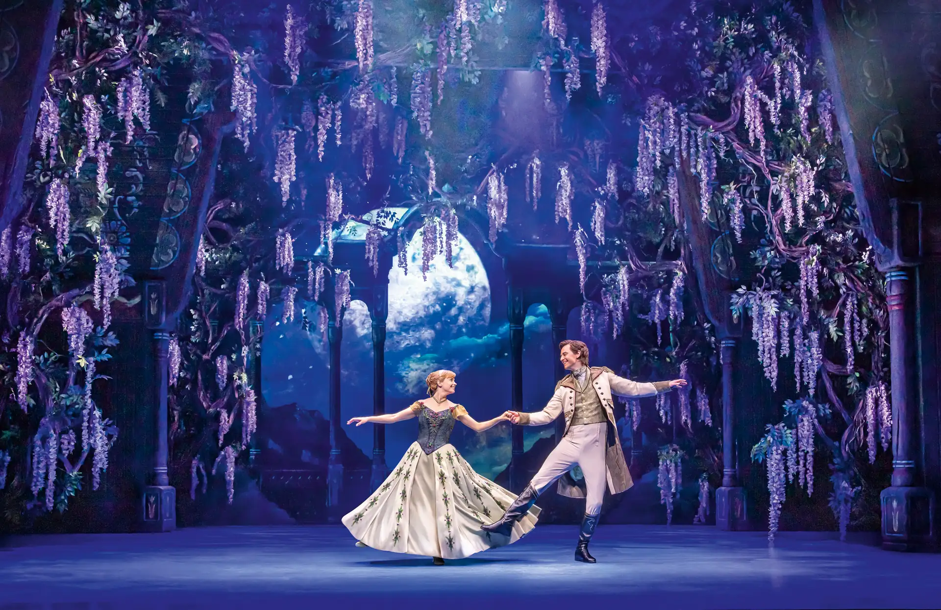 Zwei Tänzer in eleganter Kostümierung halten sich an den Händen auf einer Bühne mit hängenden Blüten und einem großen Mond im Hintergrund.