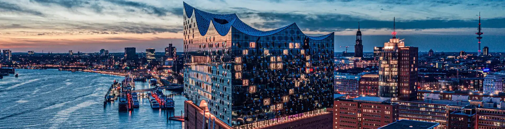 Blick auf die Elbphilharmonie bei Abenddämmerung mit beleuchteter Stadt und Hafen im Hintergrund