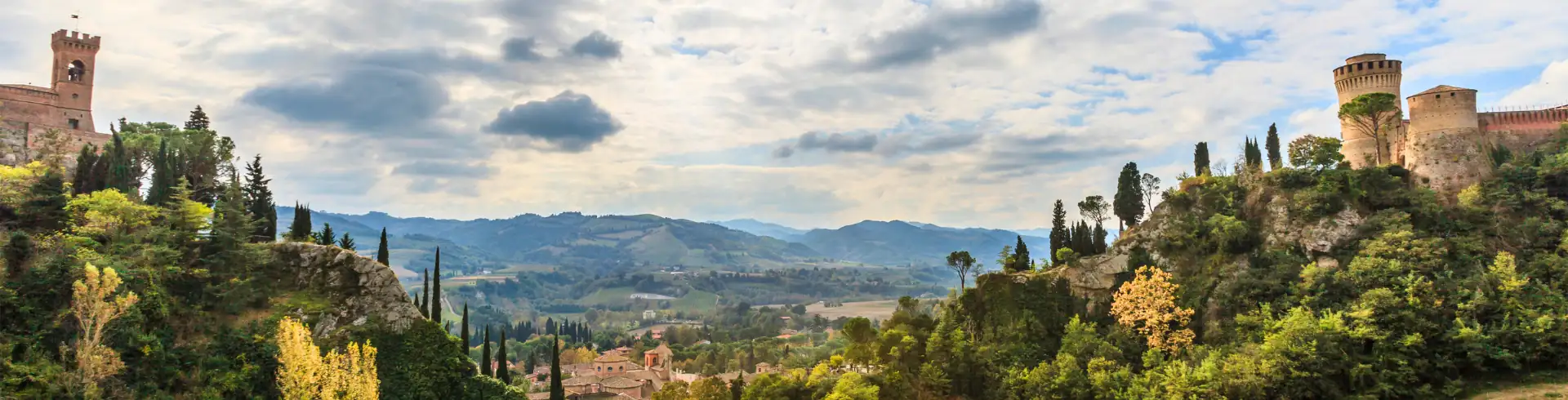 Emilia Romagna Banner: Panoramablick auf eine hügelige Landschaft bei Brisighella mit Burgen auf beiden Seiten und bewaldeten Hügeln unter bewölktem Himmel.