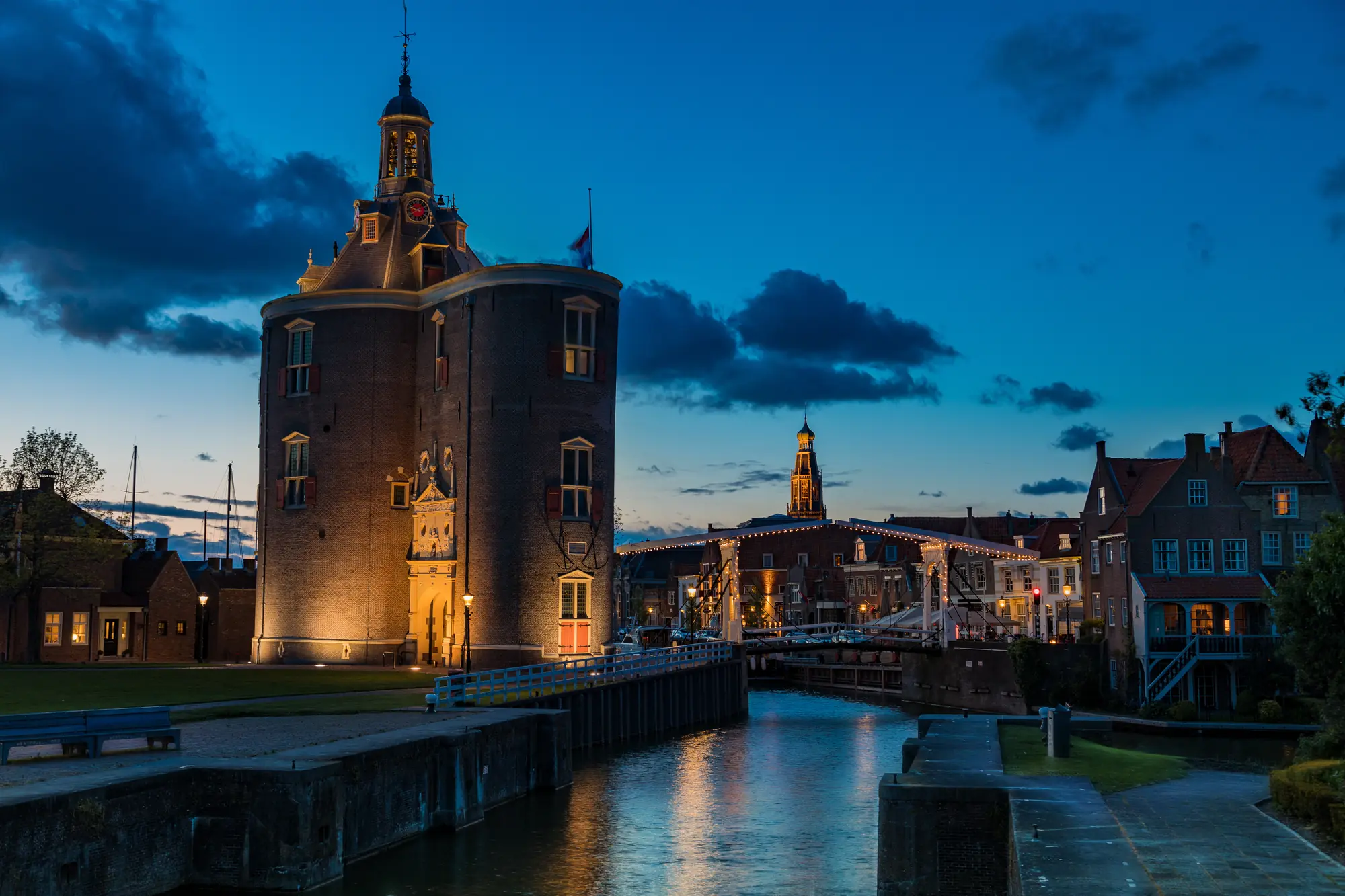 Das Drommedaris Tor in Enkhuizen zur blauen Stunde, umgeben von sanfter Stadtbeleuchtung entlang des Flusses. Die Lichter spiegeln sich im Wasser und schaffen eine malerische Atmosphäre, während der tiefblaue Himmel die historische Architektur des Tores eindrucksvoll in Szene setzt. Diese nächtliche Szenerie vermittelt ein Gefühl von Ruhe und zeitloser Schönheit.