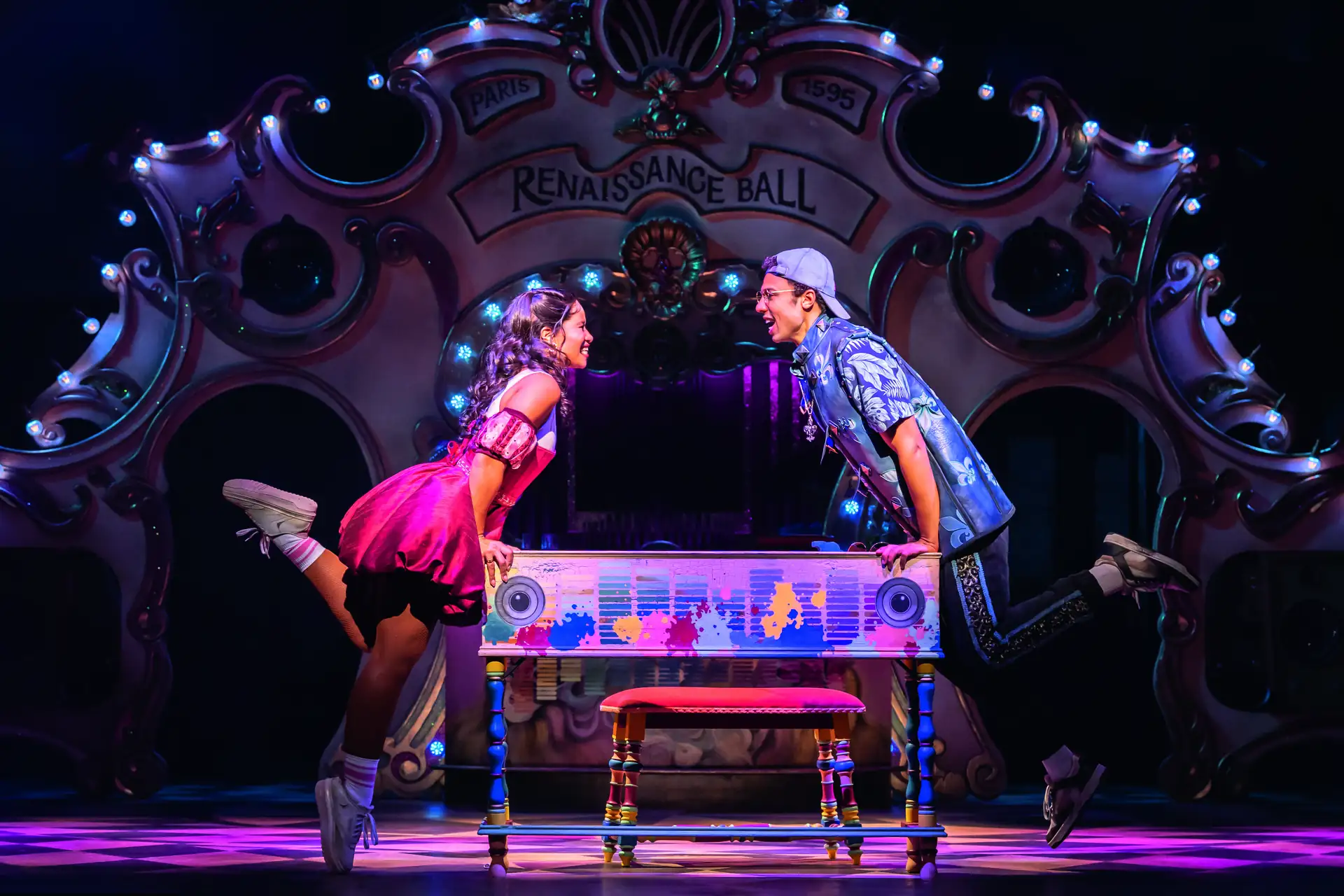 Zwei Darsteller in farbenfroher Kleidung lehnen sich auf einem bunt bemalten Klavier zueinander, im Hintergrund ein dekorativer Bogen mit der Aufschrift 'Renaissance Ball'.