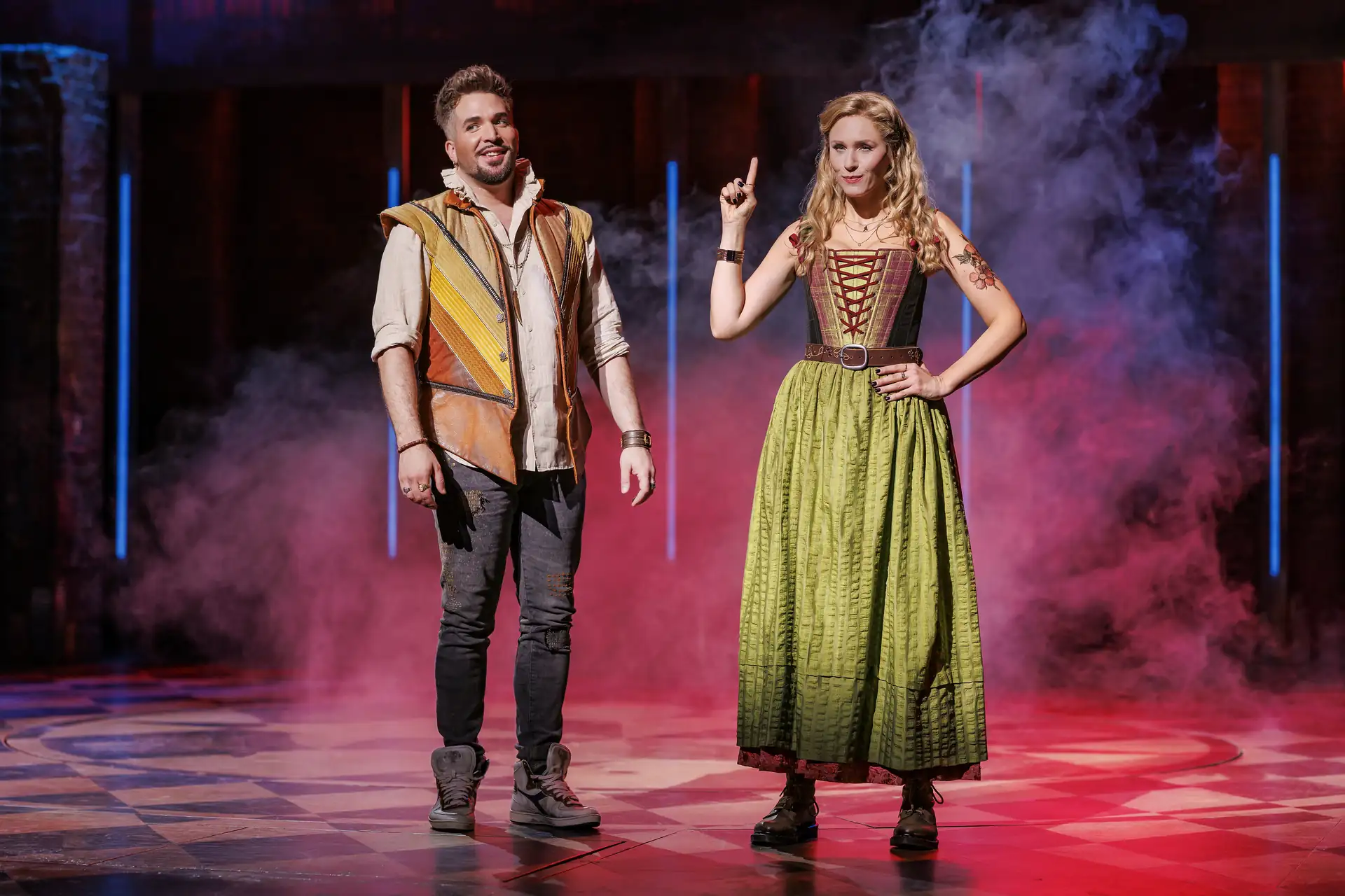 Zwei Personen auf einer Bühne mit Schachbrettmuster am Boden und rotem Nebel im Hintergrund, die Frau trägt ein langes grünes Kleid mit Schnürung, der Mann eine bunte Weste über weißem Hemd und dunkle Hosen.