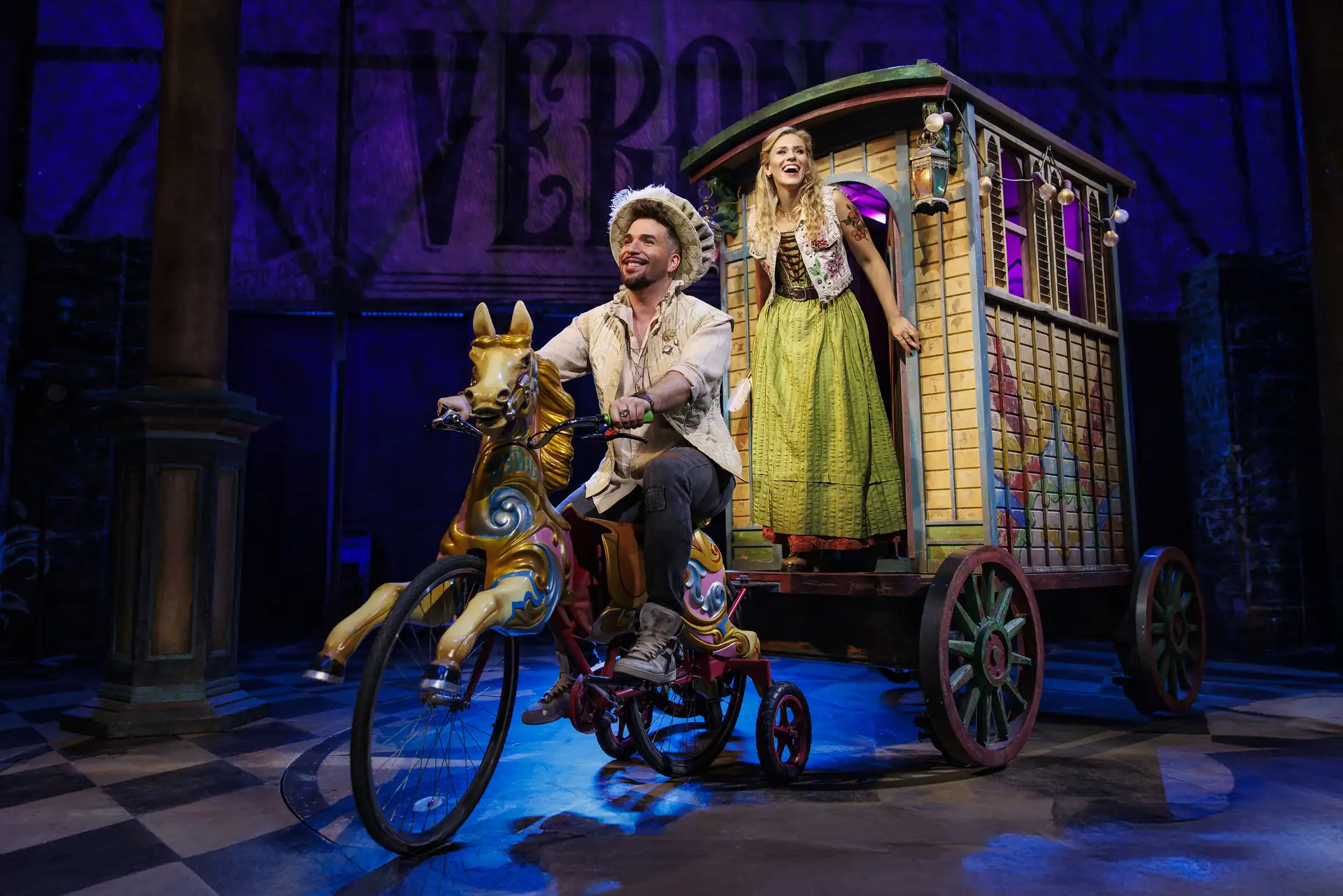 Eine Person sitzt auf einem fantasievollen, pferdeförmigen Dreirad vor einem hölzernen, rollenden Wagen, aus dem eine weitere Person in einem historischen Kleid herausschaut, im Bühnenbild mit dem Schriftzug 'VERONA' im Hintergrund.