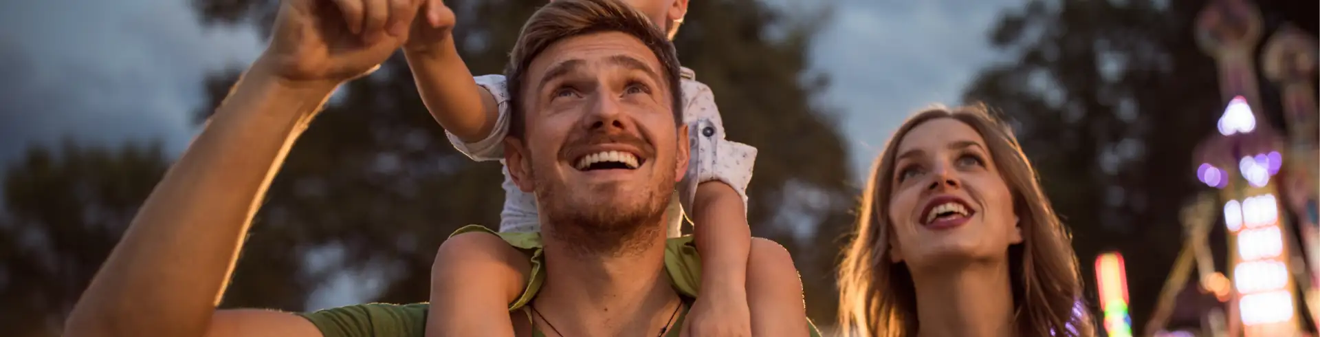 Event- und Erlebnisreisen Symbolbild im Bannerformat: Vater mit Kind auf den Schultern und Mutter daneben blicken staunend hoch, im Hintergrund sind verschwommene Lichter einer Open Air-Veranstaltung.
