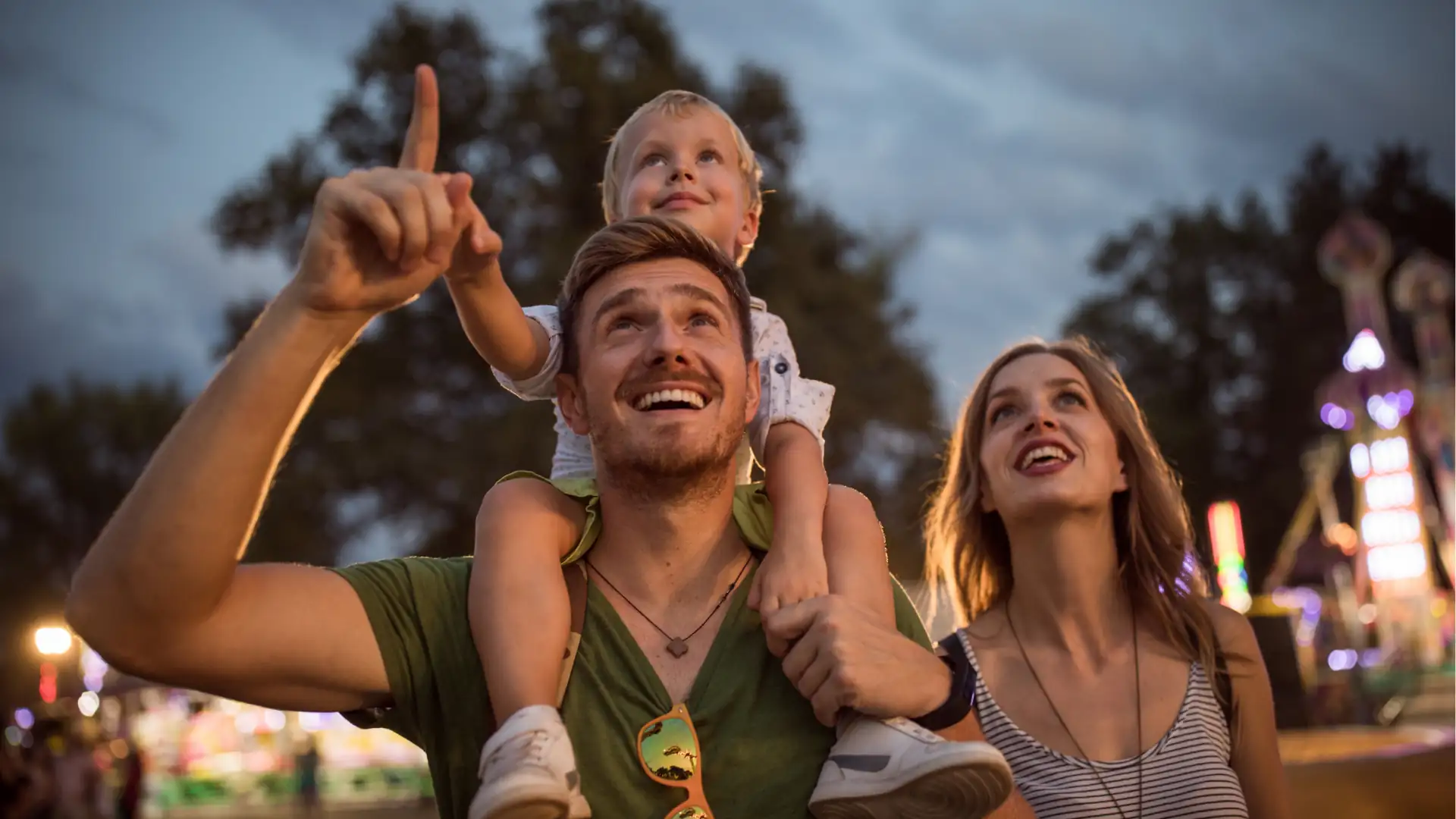 Event- und Erlebnisreisen Symbolbild: Vater mit Kind auf den Schultern und Mutter daneben blicken staunend hoch, im Hintergrund sind verschwommene Lichter einer Open Air-Veranstaltung.