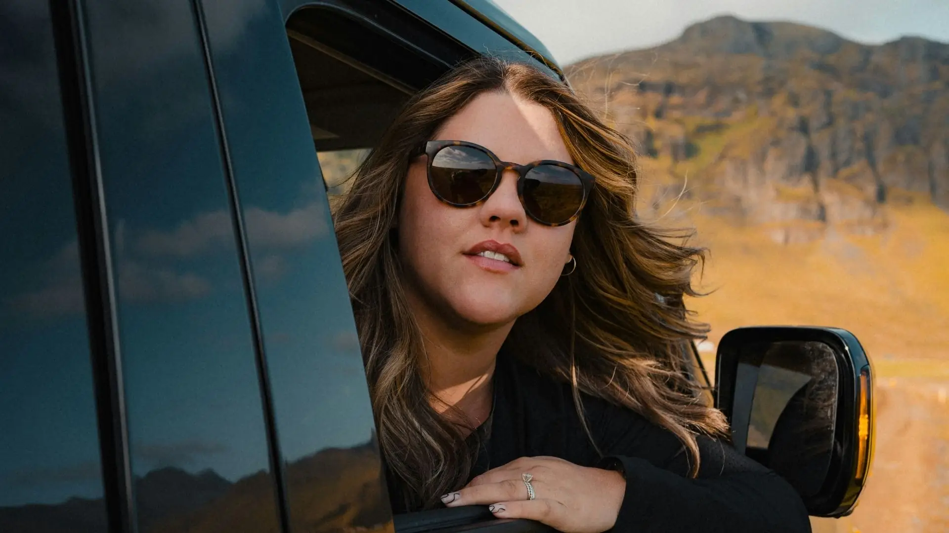 Frau lehnt aus dem Fenster eines schwarzen Autos, im Hintergrund Berge und Himmel bei Tageslicht