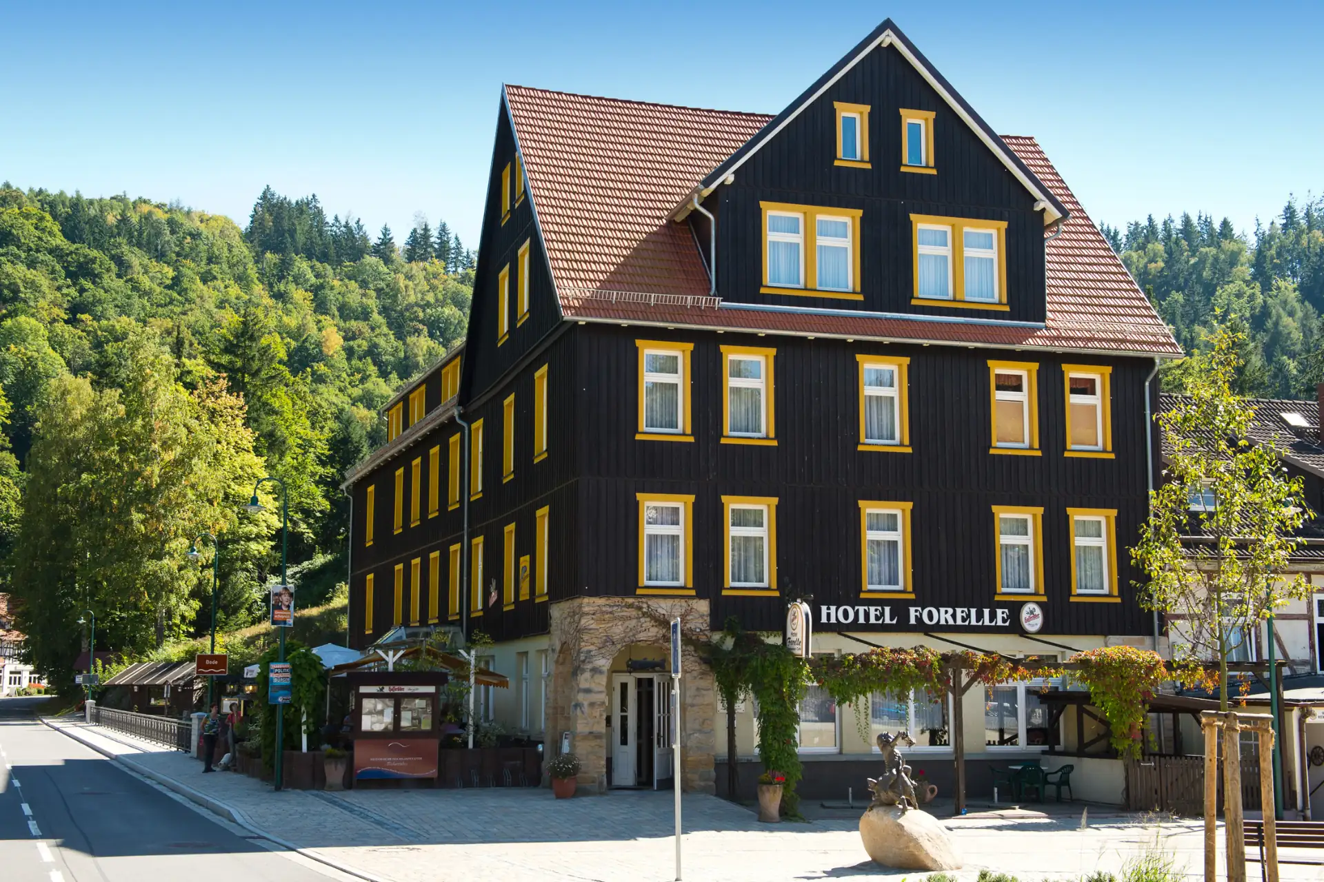 Ferienhotel Forelle Treseburg: Außenansicht des Hotels mit dunkler Holzvertäfelung, gelben Fensterrahmen und Wald im Hintergrund.