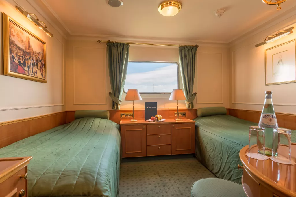 Kabine mit zwei Einzelbetten, Nachttisch mit Obst und Buch vor einem Fenster mit grünen Vorhängen, Tisch mit Wasserflasche und Gläsern