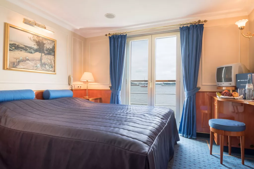 Hotelzimmer mit großem Bett, blauen Vorhängen und Blick auf Wasser durch die Balkontür