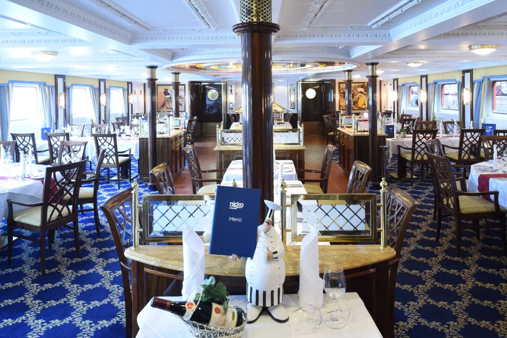 Innenraum eines Restaurants mit blauem Teppich, weißen Tischdecken und einem zentralen Holzbalken mit einem Menü und einer Dekofigur in Form eines Kochs