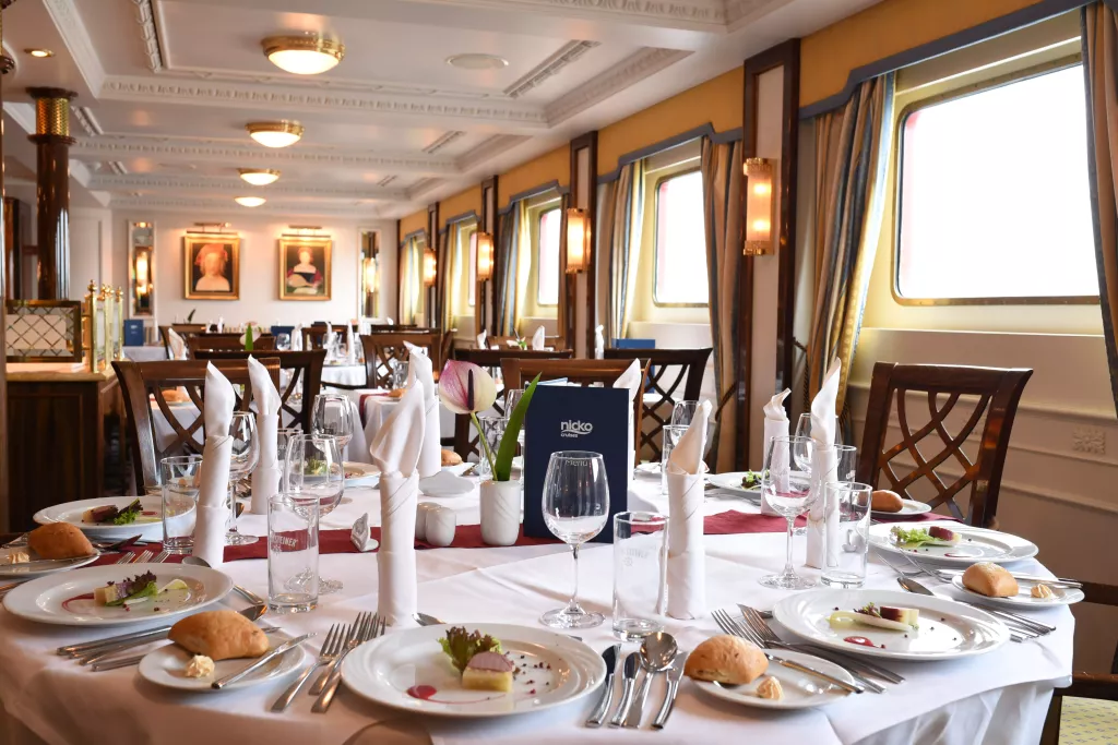 Elegantes Restaurant mit weiß gedeckten Tischen, gefalteten Servietten, Besteck und Gläsern vor großen Fenstern mit Ausblick