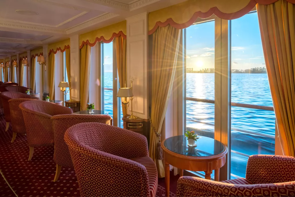 Salon mit gepolsterten Sesseln und kleinen Tischen vor großen Fenstern mit Blick auf einen Fluss oder See bei Sonnenuntergang