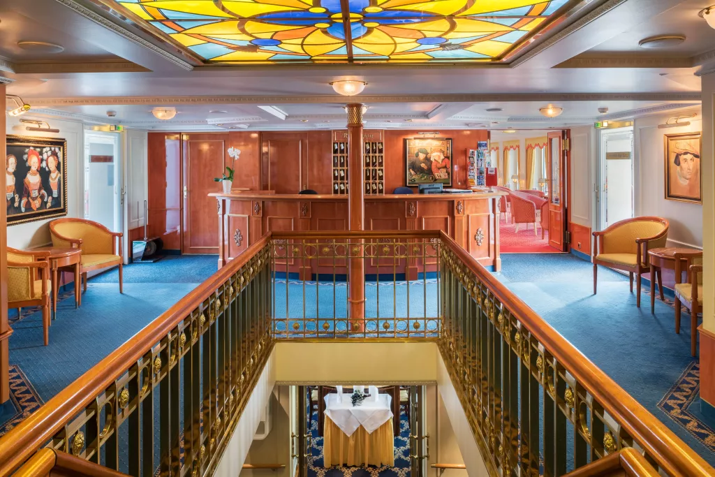 Innenansicht einer Rezeption mit blauem Teppich, goldfarbenem Geländer, Holzvertäfelung und buntem Glasfenster an der Decke