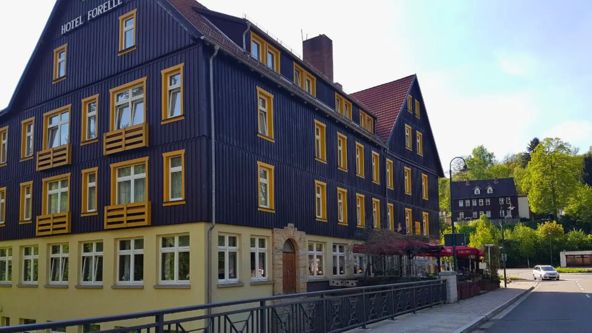 Hotel Forelle, dreistöckiges Gebäude mit dunkler Fassade und gelben Fensterrahmen an einer Straße mit Brücke