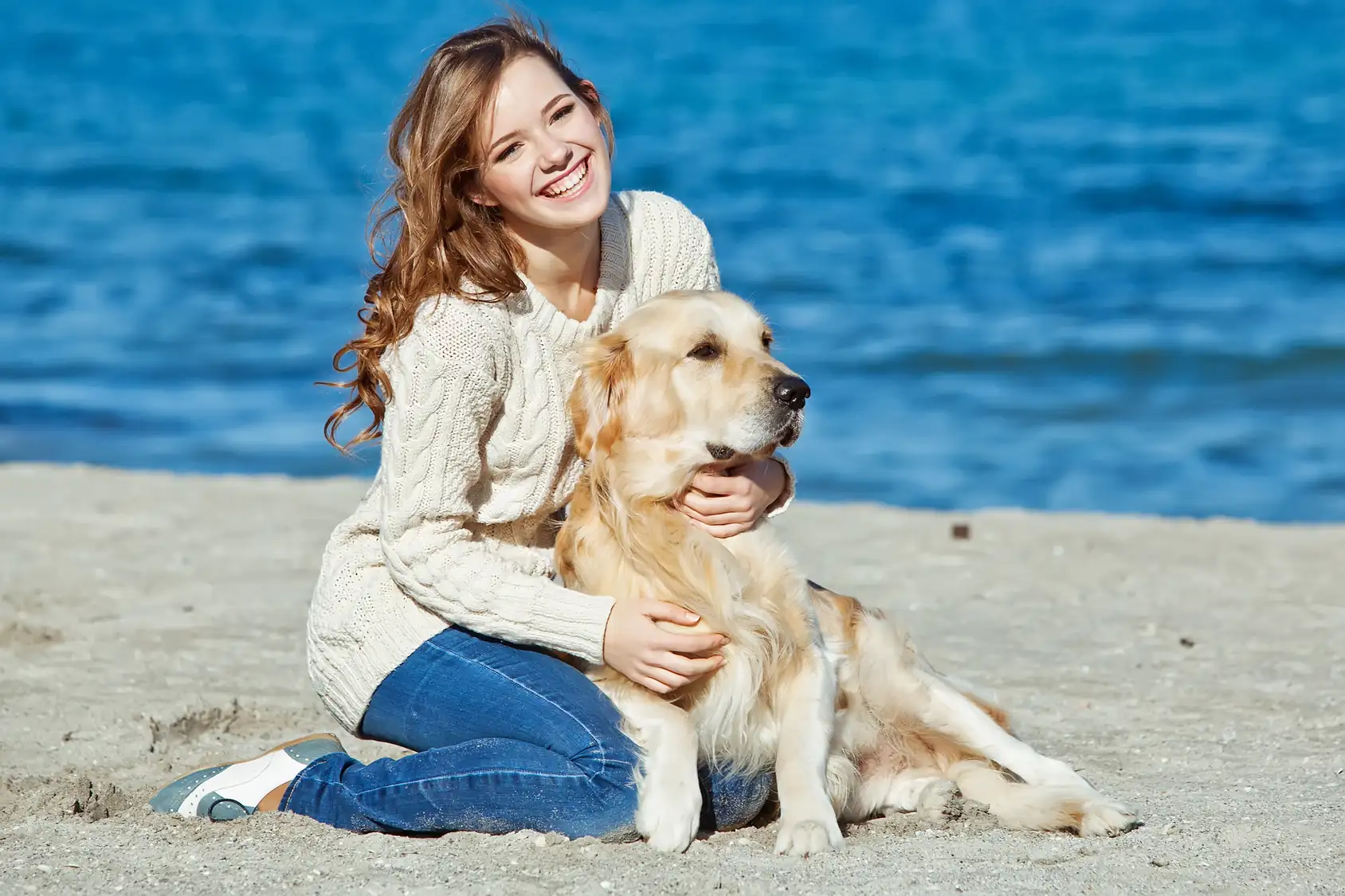 Frau sitzt am Strand und hält einen liegenden Golden Retriever im Arm