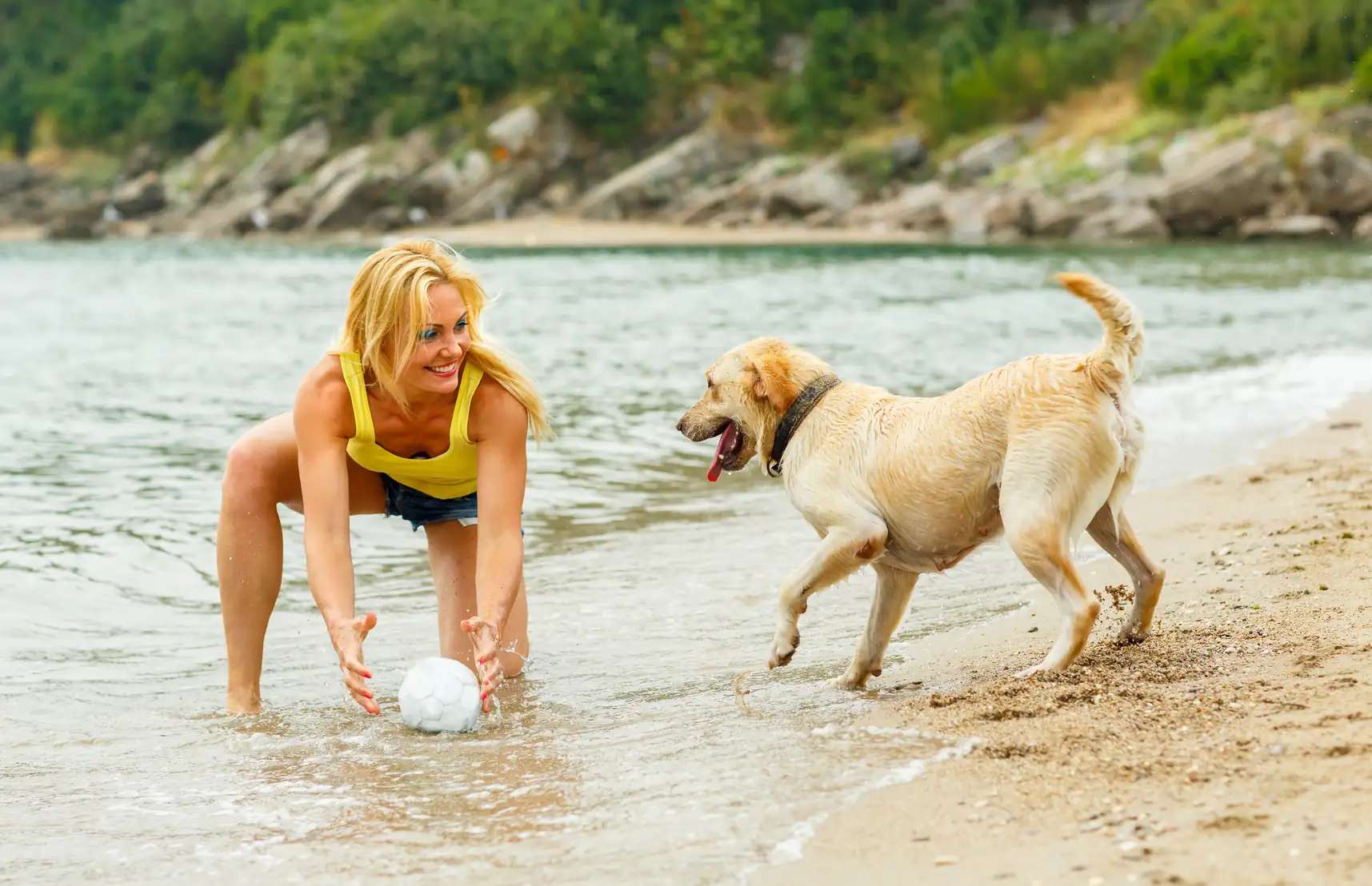 Frau in gelbem Top und Jeansshorts spielt mit hellem Hund am Ufer eines Sees oder Meeres