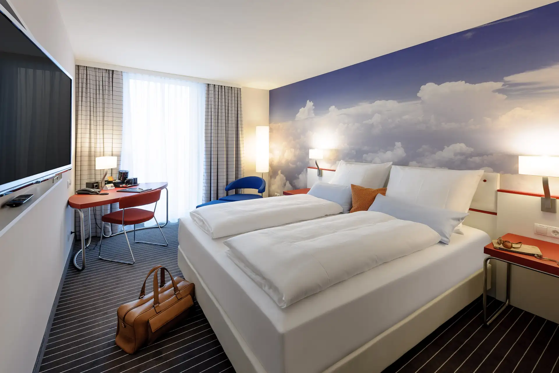 Modernes Hotelzimmer mit Doppelbett, Wandbild mit Wolkenmotiv, Schreibtisch mit Stuhl und großem Fenster mit Vorhängen im Styles Hotel in Friedrichshafen, Beispielhotel für die Radreise Bodensee