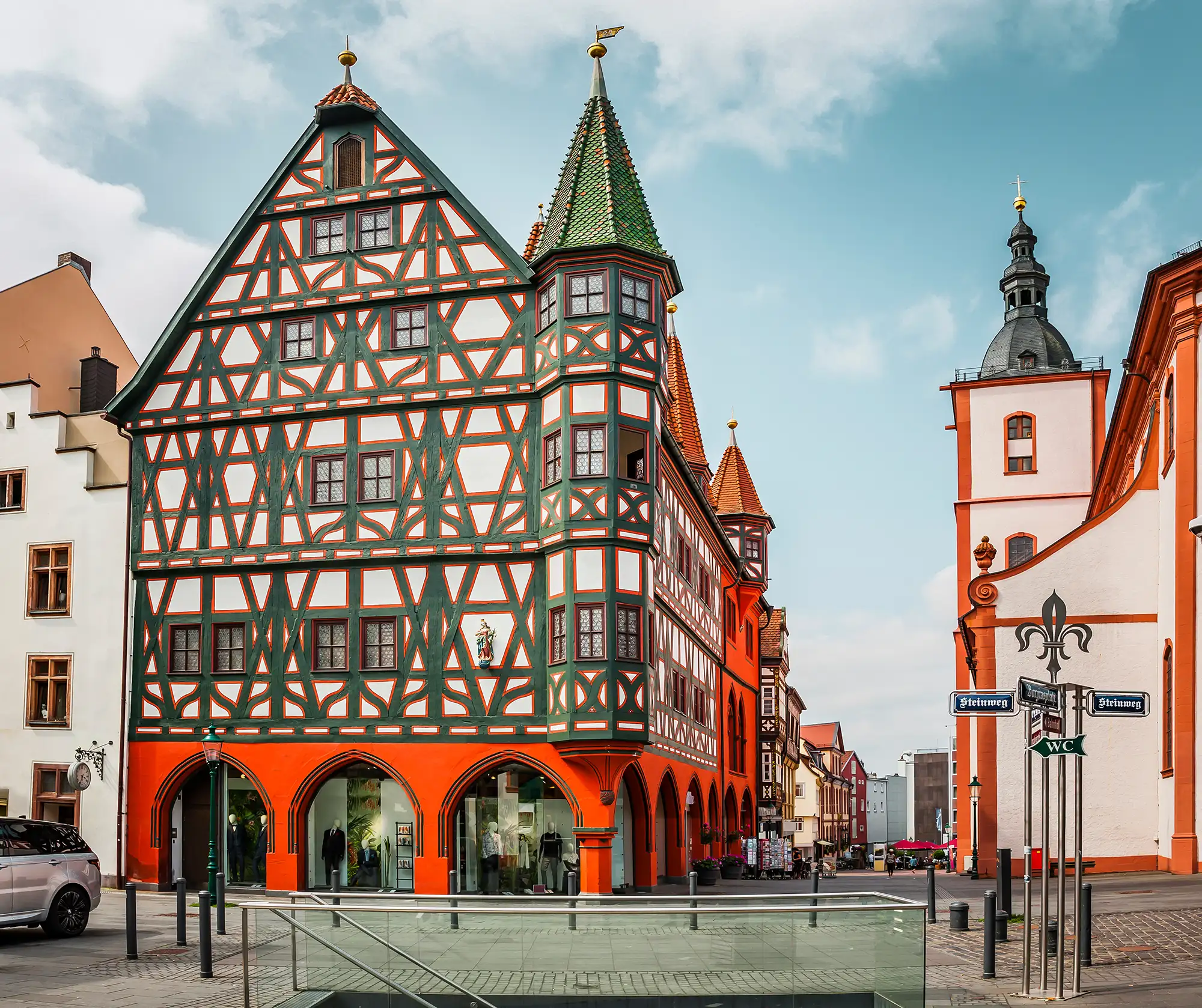 Fulda Hotel - Altes Rathaus in Fulda mit roten Bogengängen und grünen Holzbalken auf weißem Hintergrund auf einem Platz davor eine Treppe und ein Auto