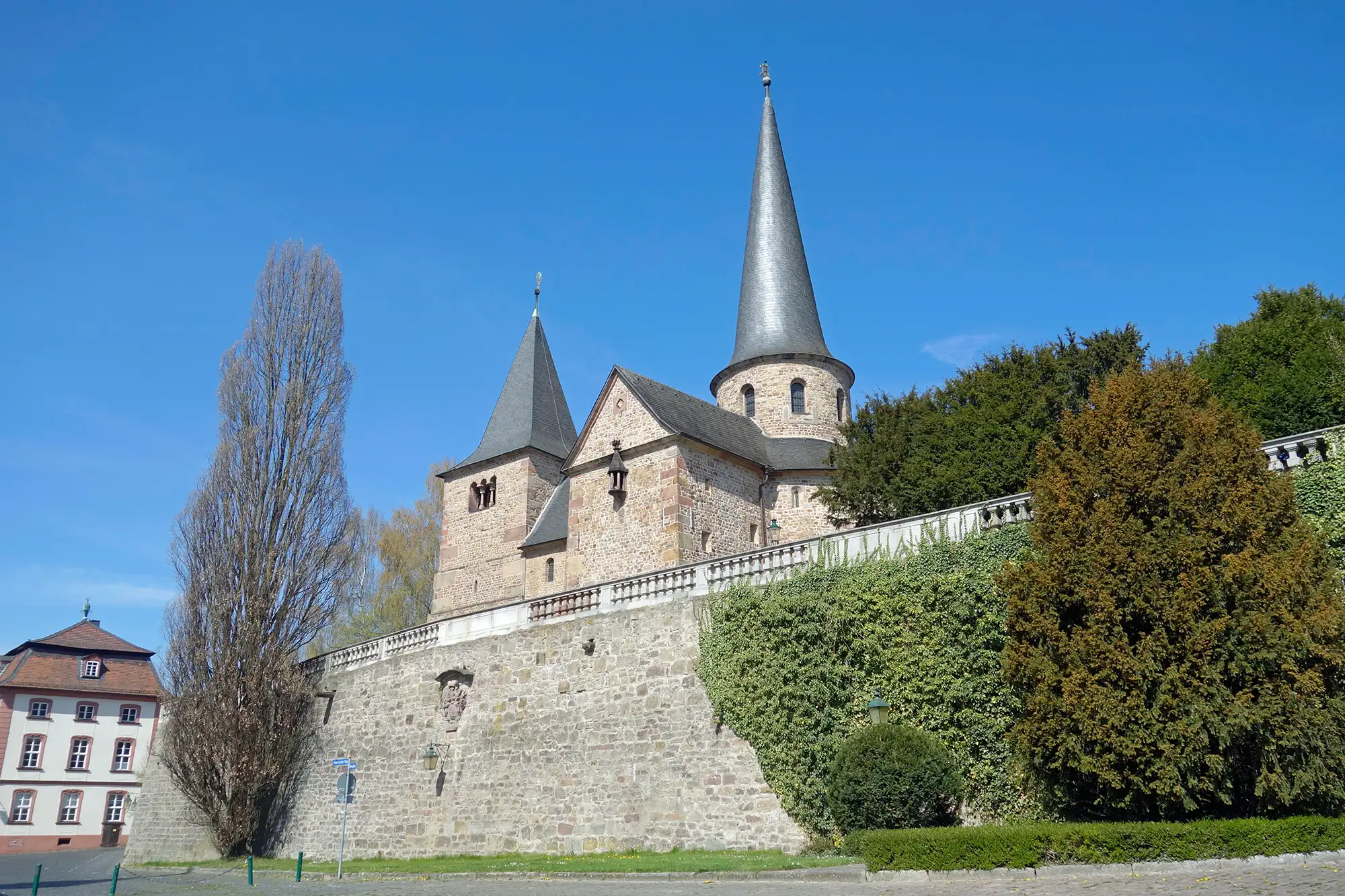 Fulda Hotel - Michaeliskirche in Fulda mit spitzen Türmen auf einer Steinmauer mit Bäumen und Büschen daneben