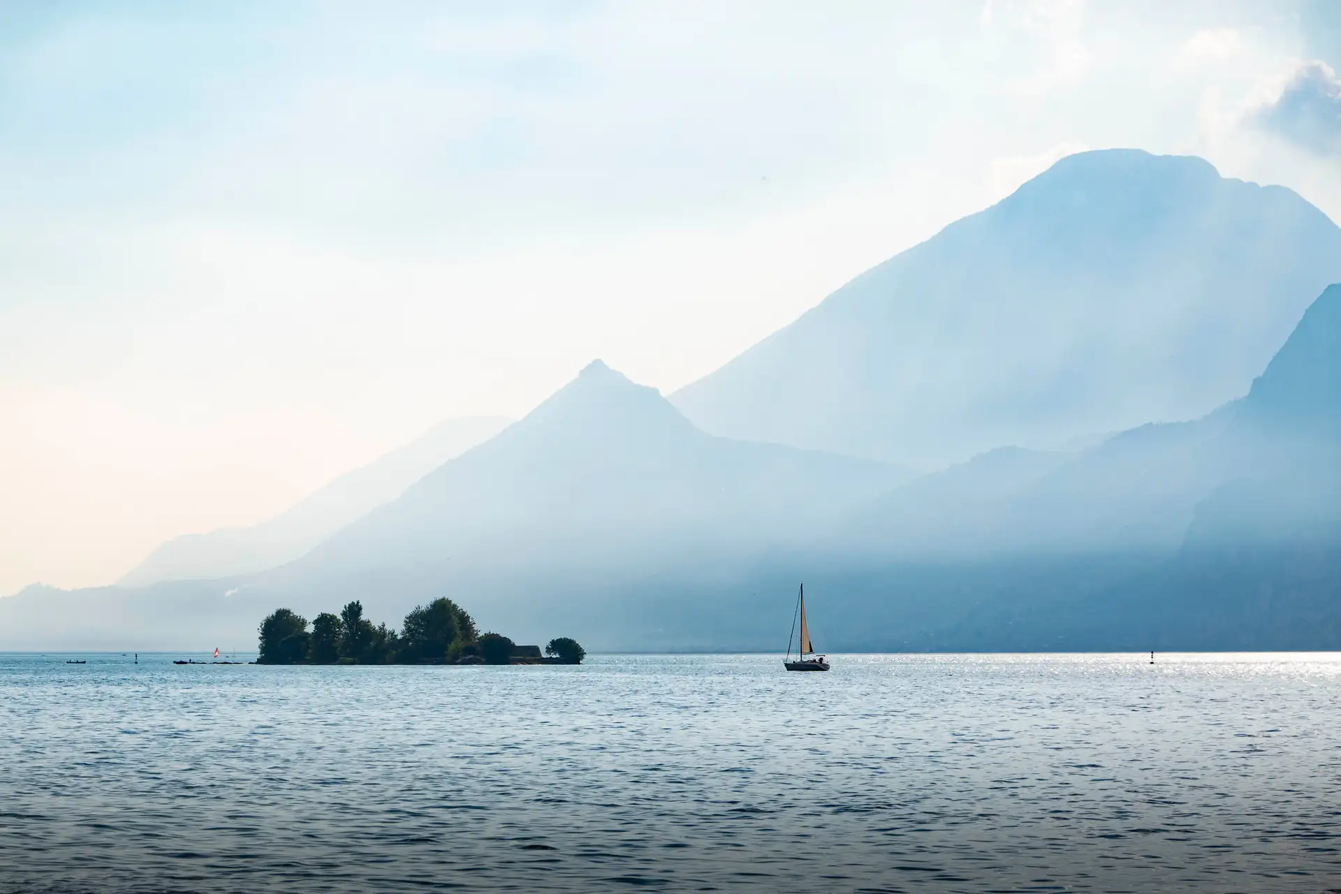 Kleine Insel namens Isola di Trimelone mit Bäumen im See vor nebelverhangenen Bergen und Segelboot auf dem Wasser