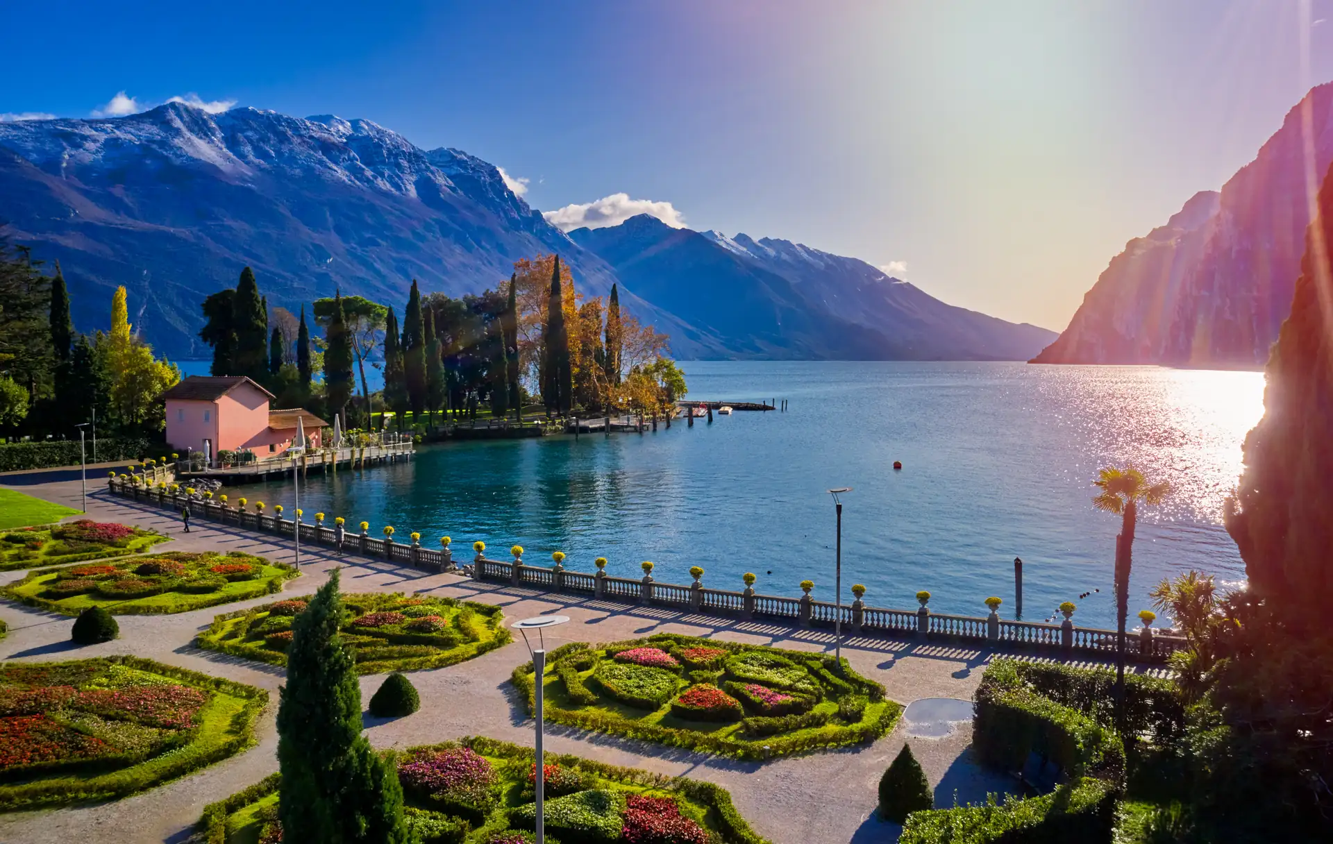Park mit symmetrischen Blumenbeeten und Palmen am Ufer des Gardasees mit Blick auf Berge und Wasser bei Sonnenlicht in Riva del Garda