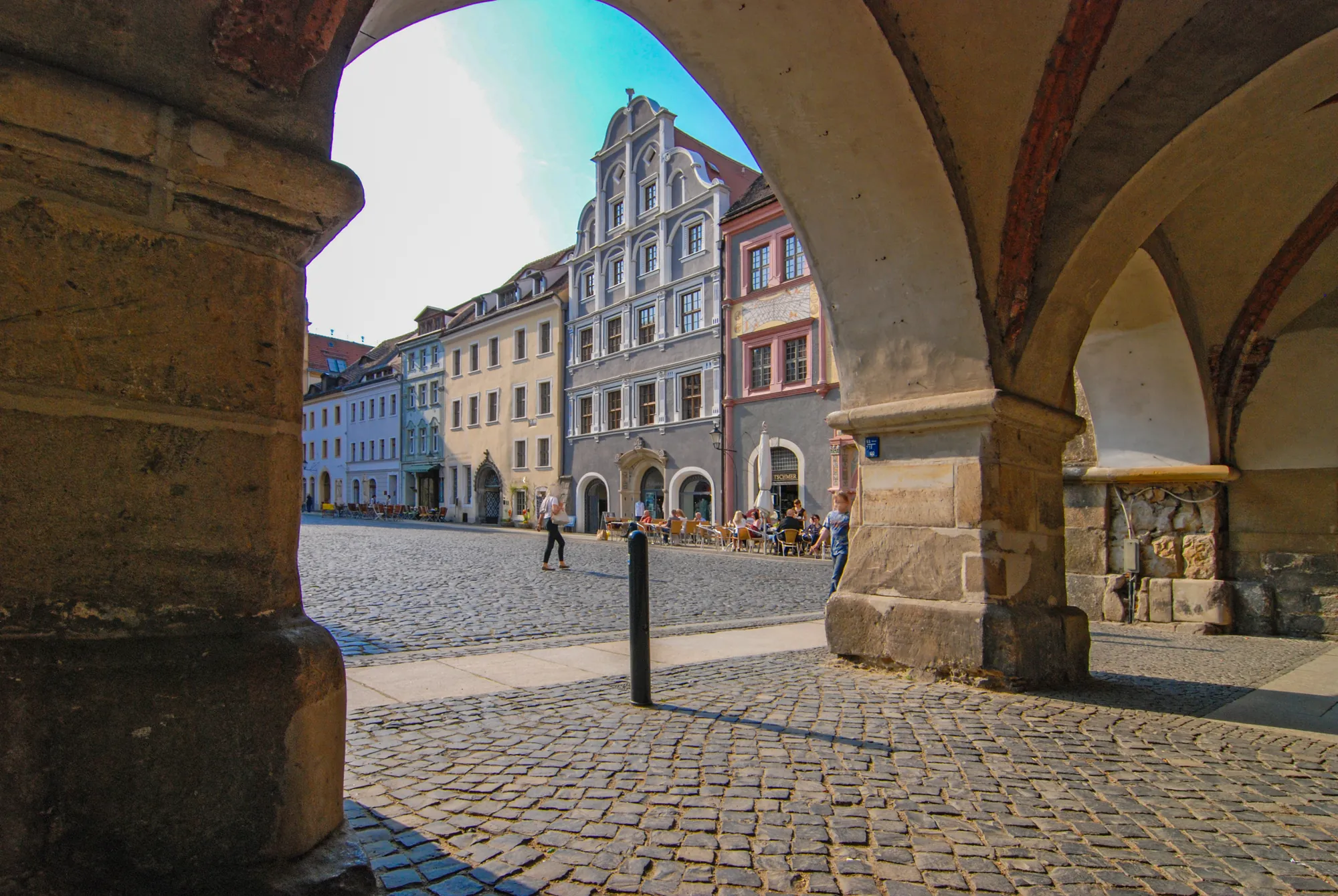 Blick auf den Untermarkt in Görlitz durch einen Bogen mit Kopfsteinpflaster und Häuserfassaden mit Menschen und Stühlen davor
