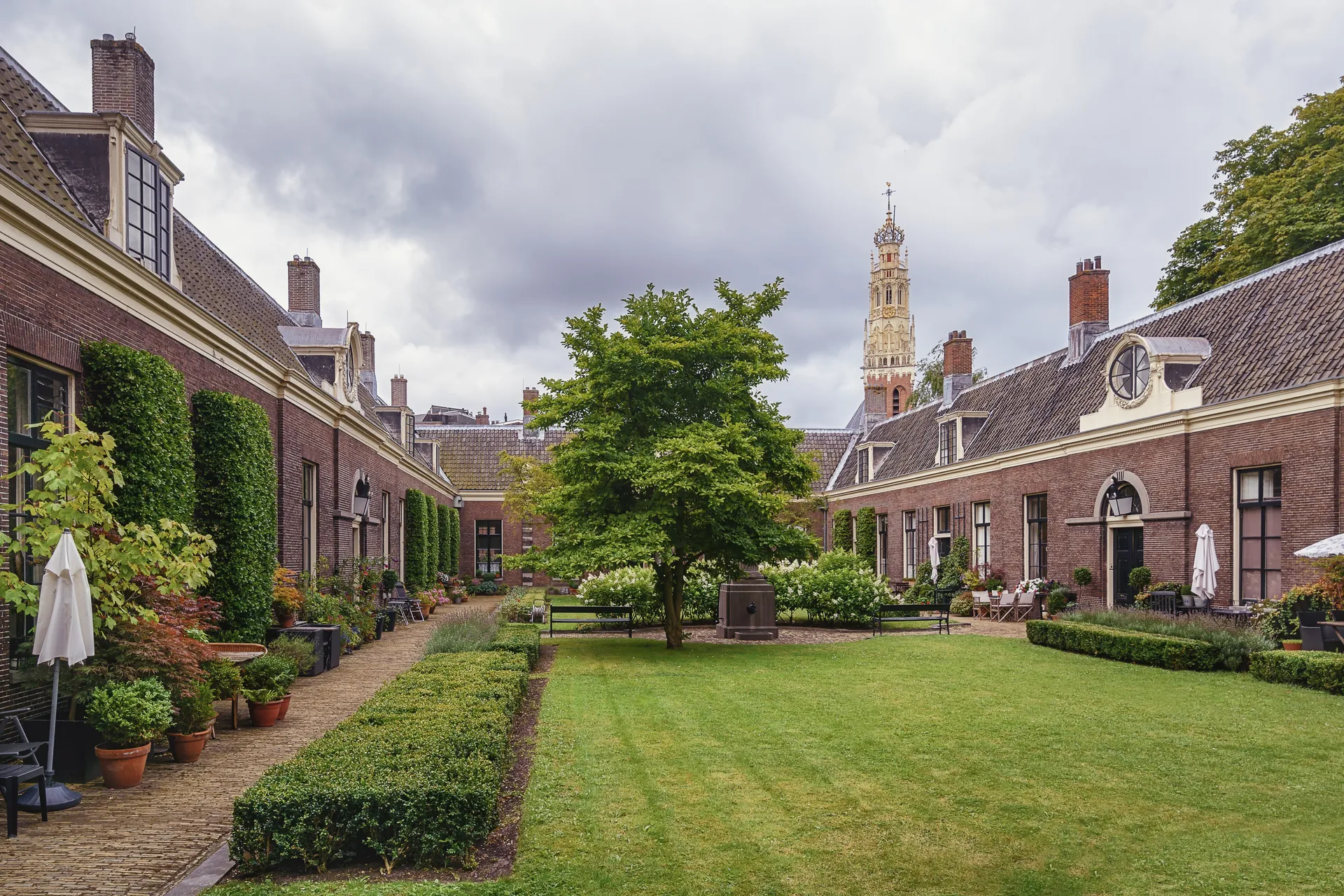Die Armenhäuser mit Hofjes in Haarlem, umgeben von einem schön gepflegten Garten, mit dem Turm der Grote Kerk (St. Bavo) im Hintergrund. Die harmonische Verbindung von historischer Architektur und üppiger Natur schafft eine einladende Atmosphäre. Diese Szenerie zeigt das kulturelle Erbe und die idyllische Schönheit der Stadt.