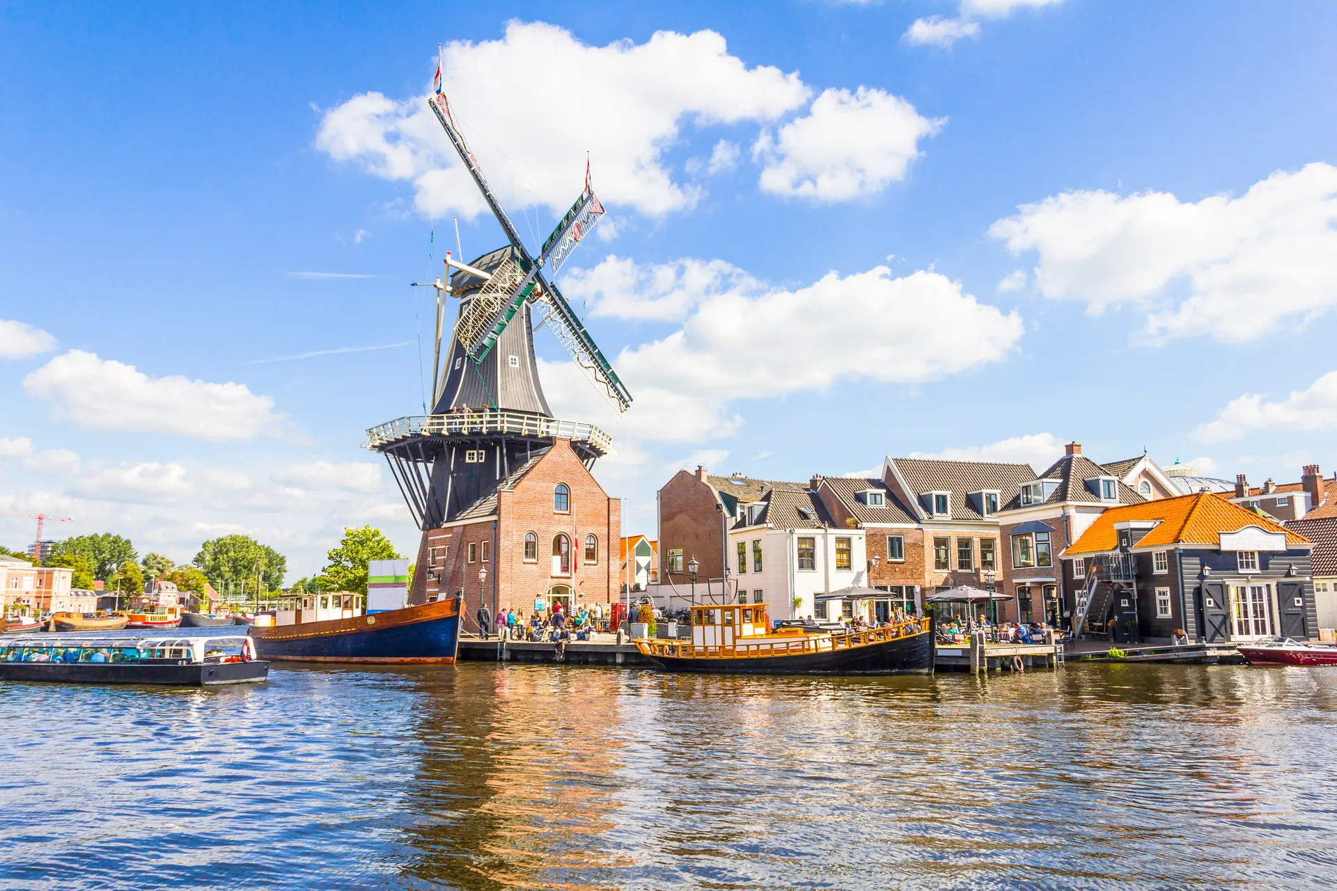 Die De Adriaan Windmühle in Haarlem, umgeben von Besuchern, bei sonnigem Wetter. Das glänzende Wasser im Vordergrund reflektiert die Mühle und verstärkt die malerische Atmosphäre. Die Szene vereint die charmante historische Architektur der Mühle mit der lebhaften Präsenz der Menschen und der natürlichen Schönheit der Umgebung.