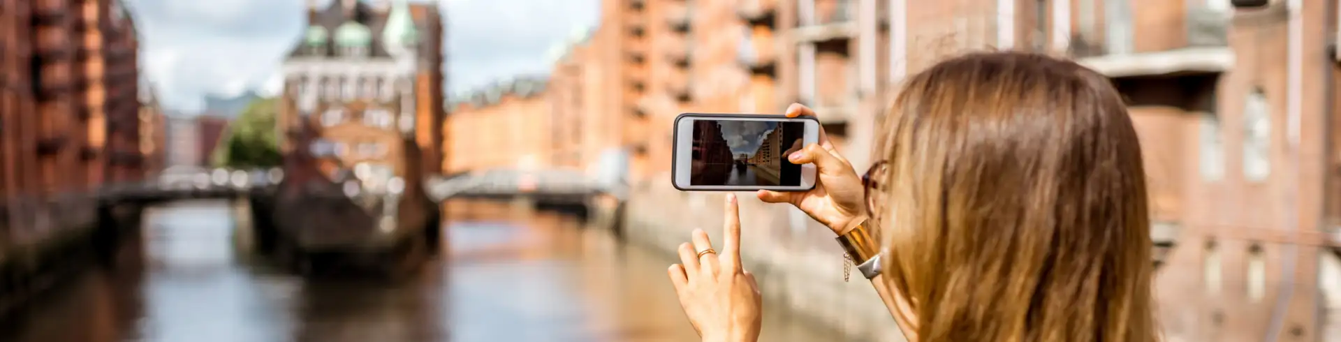 Frau mit langen braunen Haaren fotografiert mit Smartphone eine historische Speicherstadt am Wasser