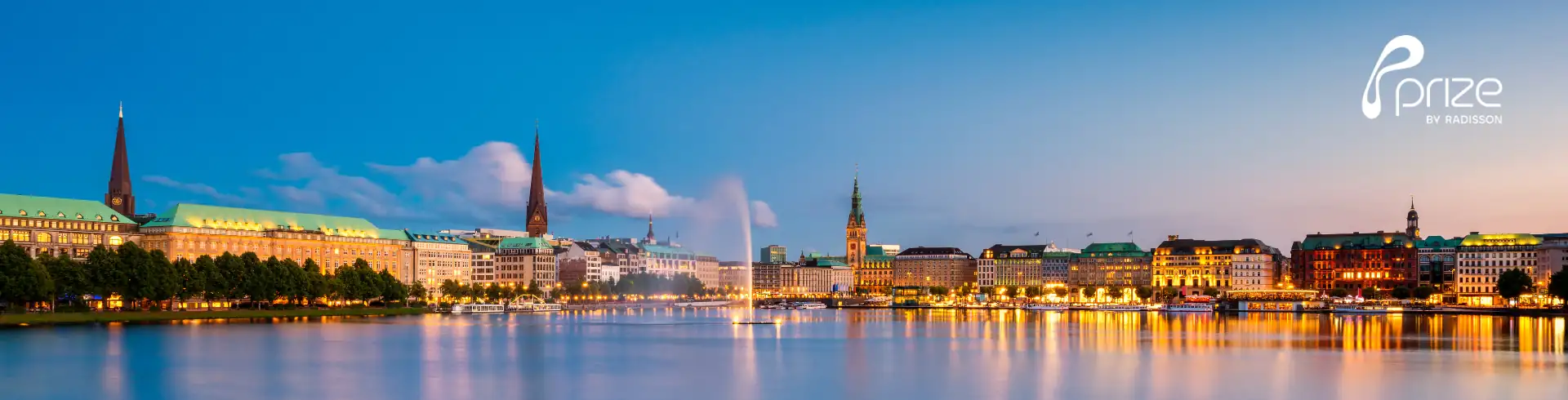 Panoramablick auf die Hamburger Binnenalster mit beleuchteten Gebäuden und zwei Kirchtürmen bei Abenddämmerung