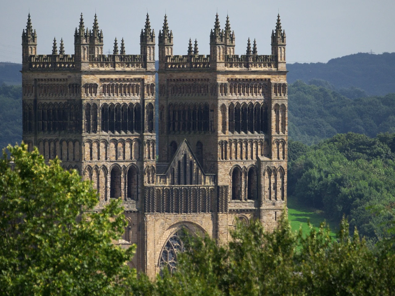 Das Bild zeigt einen beeindruckenden Blick auf die Durham Cathedral in England, eines der berühmtesten Beispiele normannischer Architektur. Die massiven Türme der Kathedrale erheben sich majestätisch über die umliegenden Bäume und Wälder. Die detaillierte Gestaltung der Türme, mit ihren zahlreichen Bögen und Zinnen, verleiht der Kathedrale einen imposanten und zugleich kunstvollen Charakter. Im Hintergrund sind die grünen Hügel und Wälder zu sehen, die der Szene eine natürliche und ruhige Atmosphäre verleihen, während die historische Bedeutung der Kathedrale deutlich sichtbar wird.