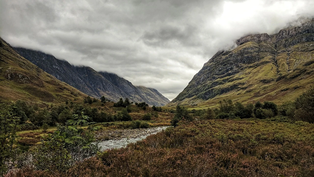 Das Bild zeigt die beeindruckende Landschaft von Glencoe, einem der spektakulärsten Täler in Schottland. Umgeben von steilen, bewaldeten Bergen, verleiht der bewölkte Himmel der Szene eine dramatische Atmosphäre. Ein schmaler Fluss schlängelt sich durch das Tal, während die üppige Vegetation und das farbenfrohe Heidekraut den Boden bedecken. Die majestätische Natur von Glencoe ist ein beliebtes Ziel für Wanderer und Naturliebhaber, die die atemberaubenden Aussichten und die ruhige, unberührte Schönheit genießen möchten.