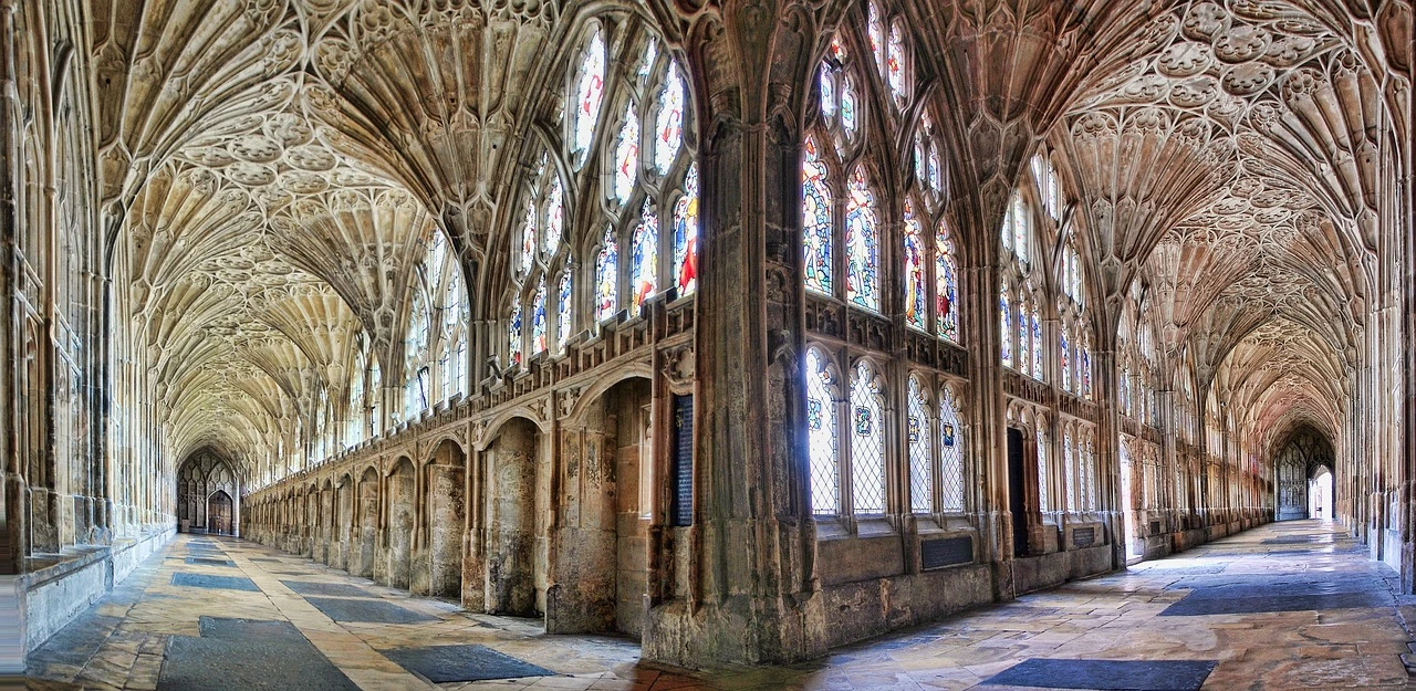 Das Bild zeigt die atemberaubenden Gewölbegänge der Gloucester Cathedral in England. Die gotischen Spitzbögen, kunstvollen Verzierungen und prächtigen Glasfenster erzeugen eine majestätische Atmosphäre. Die feinen Steinmetzarbeiten an der Decke und den Wänden verleihen der Kathedrale eine besondere Eleganz. Licht strömt durch die farbenfrohen Glasfenster und wirft bunte Reflexionen auf den Boden, was die spirituelle und architektonische Schönheit des Raums unterstreicht. Die endlos erscheinenden Gänge laden zu einer Entdeckungsreise durch die Geschichte und Architektur dieser prächtigen Kathedrale ein.