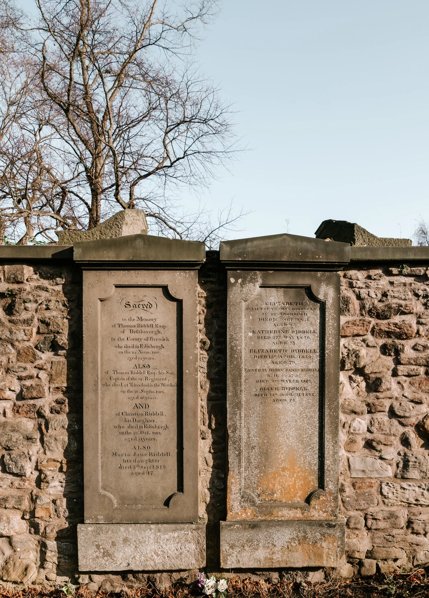 Das Bild zeigt einen Teil des Greyfriars Kirkyard in Edinburgh, einem historischen Friedhof, der für seine beeindruckenden Grabsteine und die umgebende Architektur bekannt ist. In der Mitte des Bildes sind zwei auffällige Grabsteine zu sehen, die detaillierte Inschriften aufweisen und die Namen der Verstorbenen ehren. Die Steinmauern im Hintergrund und die kahlen Bäume im oberen Bildbereich verleihen der Szene eine melancholische, aber auch ruhige Atmosphäre. Der Friedhof ist nicht nur ein Ort des Gedenkens, sondern auch ein beliebtes Ziel für Touristen, die mehr über die Geschichte und die berühmten Persönlichkeiten, die hier begraben sind, erfahren möchten.