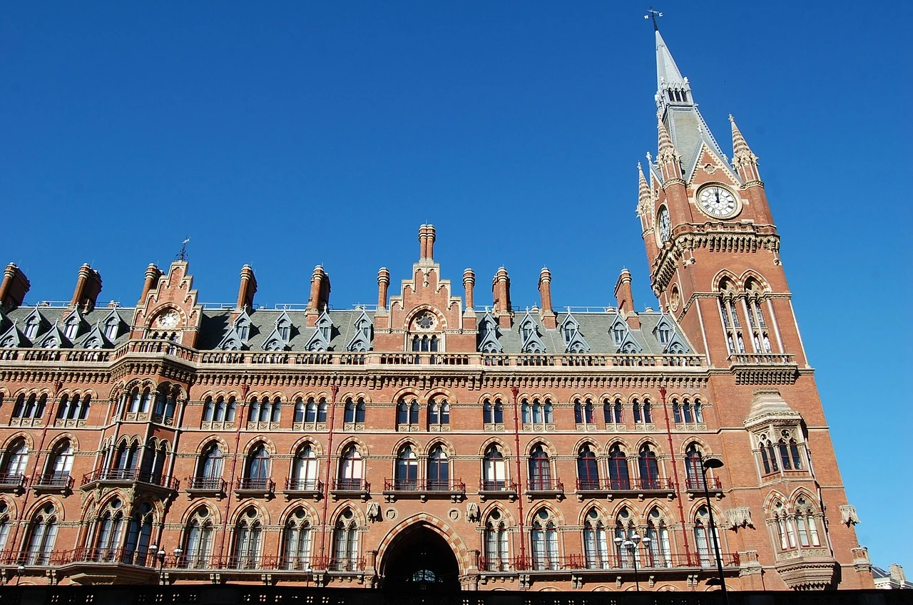 Das Bild zeigt den prächtigen Gebäudekomplex des St Pancras Bahnhofs in London. Die beeindruckende Architektur im neugotischen Stil zeichnet sich durch die roten Ziegelsteine und die zahlreichen Türmchen sowie dekorative Elemente aus. Der große Uhrturm sticht hervor, und die detaillierte Gestaltung der Fenster und Fassade unterstreicht die viktorianische Pracht dieses ikonischen Gebäudes. St Pancras ist nicht nur ein wichtiger Verkehrsknotenpunkt, sondern auch ein architektonisches Wahrzeichen Londons.