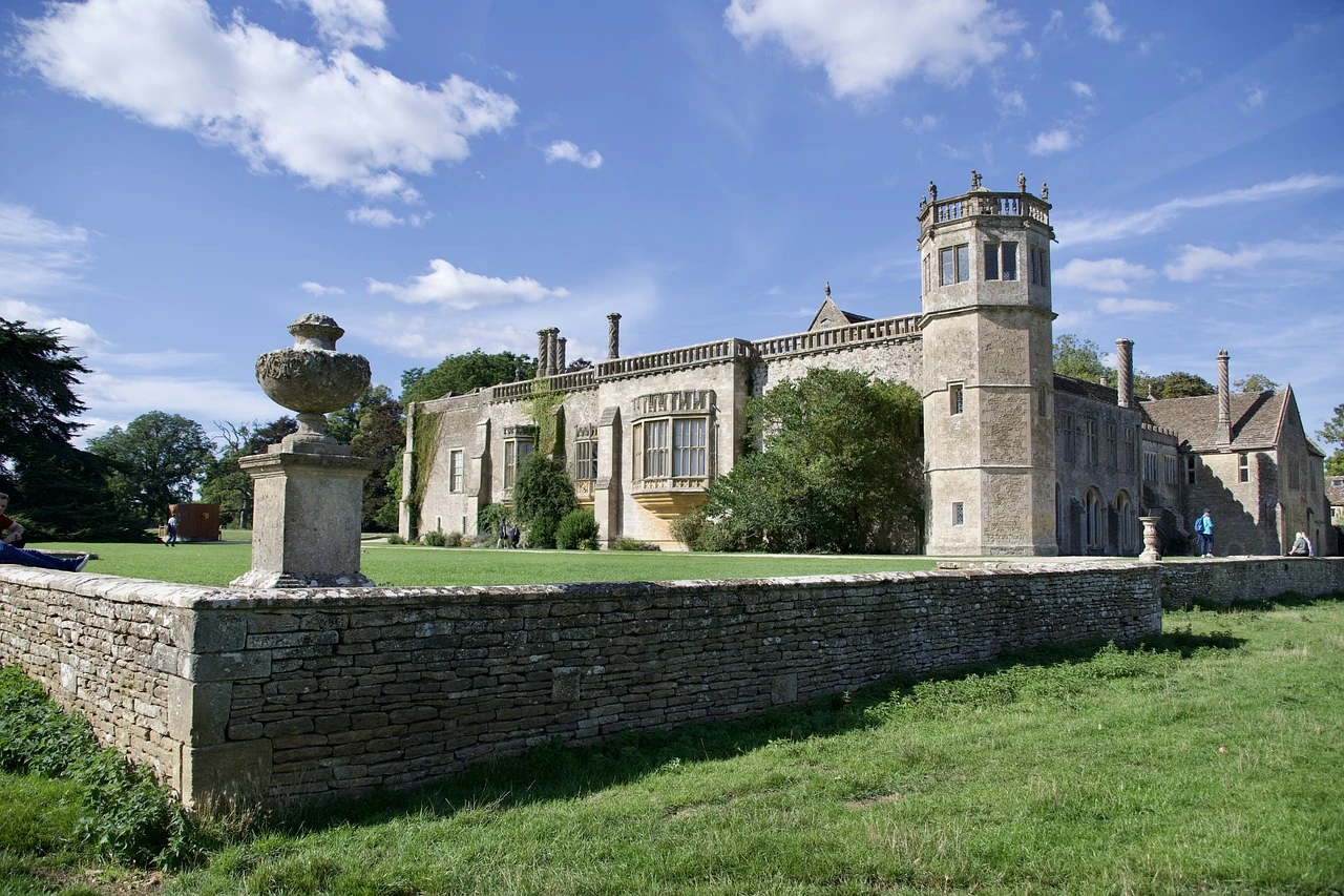 Das Bild zeigt Lacock Abbey, ein historisches Gebäude in Wiltshire, England. Die Abtei, die ursprünglich im 13. Jahrhundert als Nonnenkloster gegründet wurde, zeichnet sich durch ihre gotische Architektur aus. Auffällig sind die mit Efeu bewachsene Fassade, große Fenster sowie der markante Turm auf der rechten Seite. Eine niedrige Steinmauer verläuft entlang des Vorgartens, der von grünen Wiesen umgeben ist. Der blaue Himmel und das gepflegte Gelände verstärken den Eindruck einer ruhigen, malerischen Umgebung.