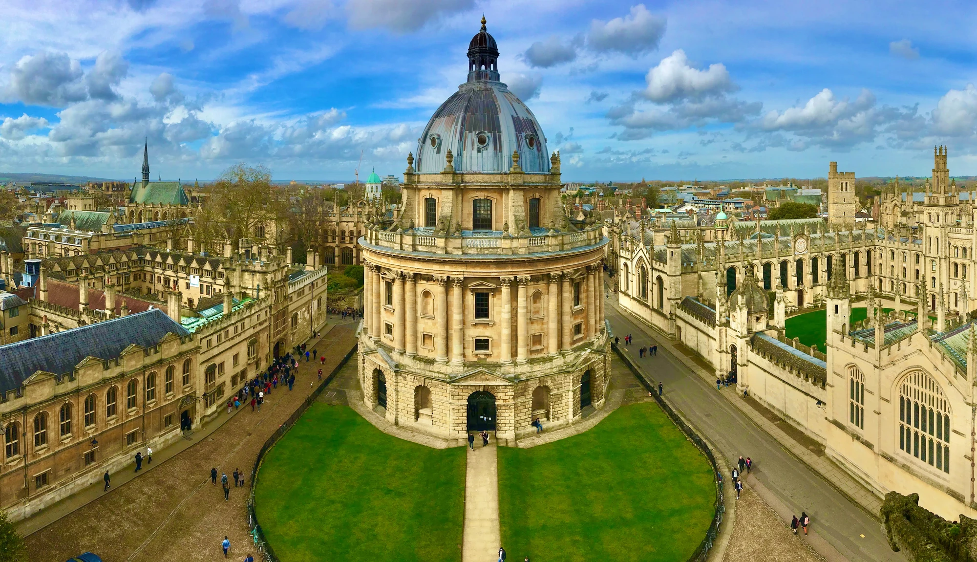Das Bild zeigt eine Panoramaansicht der University of Oxford, umgeben von historischen Gebäuden und gepflegten Gärten. Menschen schlendern gemütlich durch die weitläufigen Campusbereiche, was eine lebendige und einladende Atmosphäre schafft. Die imposante Architektur der Universität ist im Hintergrund sichtbar. Diese Szenerie verkörpert die Verbindung von Bildung, Geschichte und Gemeinschaft und lädt den Betrachter ein, die Schönheit und den Geist des akademischen Lebens in Oxford zu erleben.