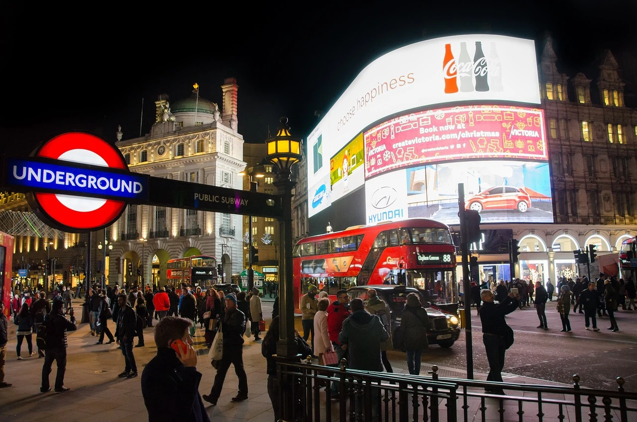 Das Bild zeigt eine lebhafte Szene bei Nacht in London, wahrscheinlich am Piccadilly Circus, einem der bekanntesten Orte der Stadt. Im Vordergrund befindet sich ein leuchtendes Schild der Londoner U-Bahn (Underground), das den Eingang zur U-Bahn-Station markiert. Menschen strömen durch die Straßen, während im Hintergrund große, farbenfrohe digitale Werbetafeln leuchten, die berühmte Marken und Anzeigen zeigen. Ein ikonischer roter Doppeldeckerbus fährt an der Menge vorbei, was die typische Londoner Atmosphäre unterstreicht. Die historischen Gebäude umgeben die Szene, die von den hellen Lichtern und der geschäftigen Aktivität geprägt ist.