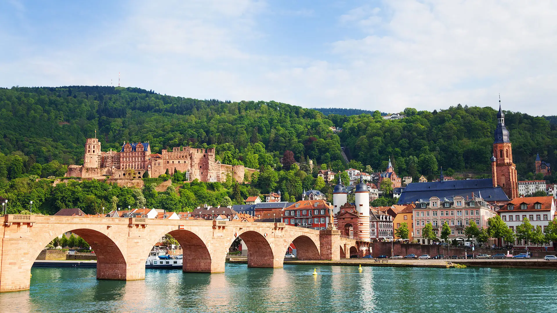 Blick auf die Alte Brücke über den Neckar mit dem Schloss Heidelberg und bewaldeten Hügeln im Hintergrund