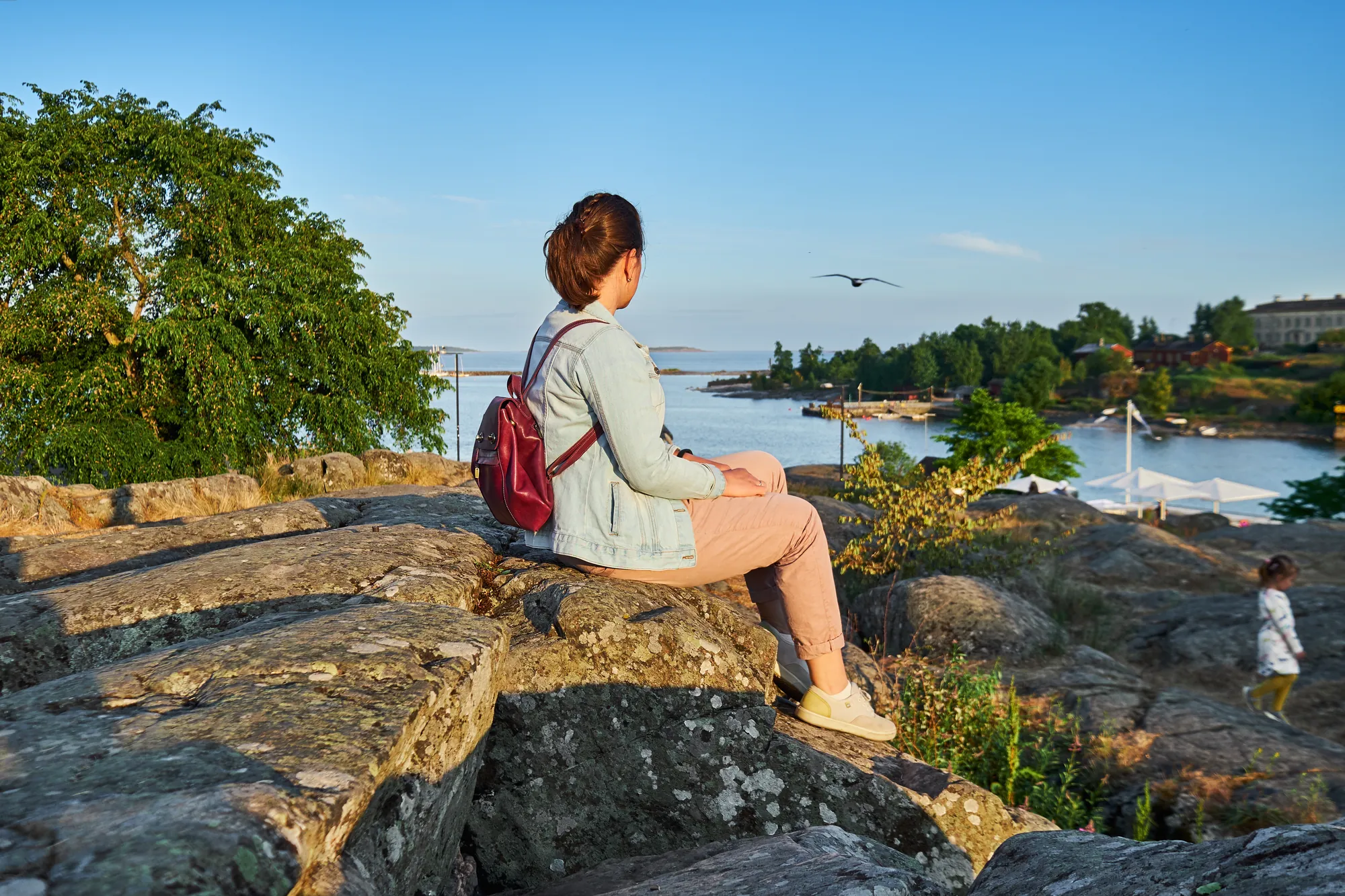 Frau sitzt auf einem Felsen im Kaivopuisto Park in Helsinki und betrachtet die atemberaubende Natur mit dem Wasser, umgeben von einer ruhigen und malerischen Landschaft.
