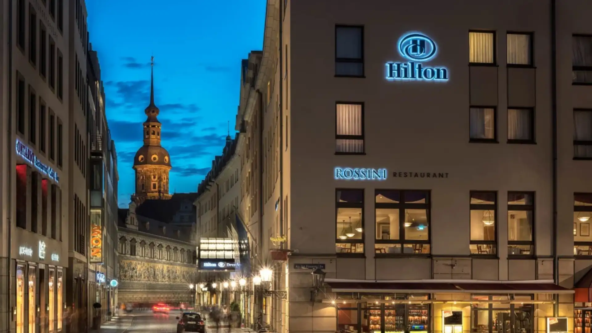 Hilton Hotels im Kachelformat: Hilton Dresden mit beleuchtetem Logo und Restaurant 'Rossini' in einem mehrstöckigen Gebäude bei Abenddämmerung in einer Altstadtstraße.