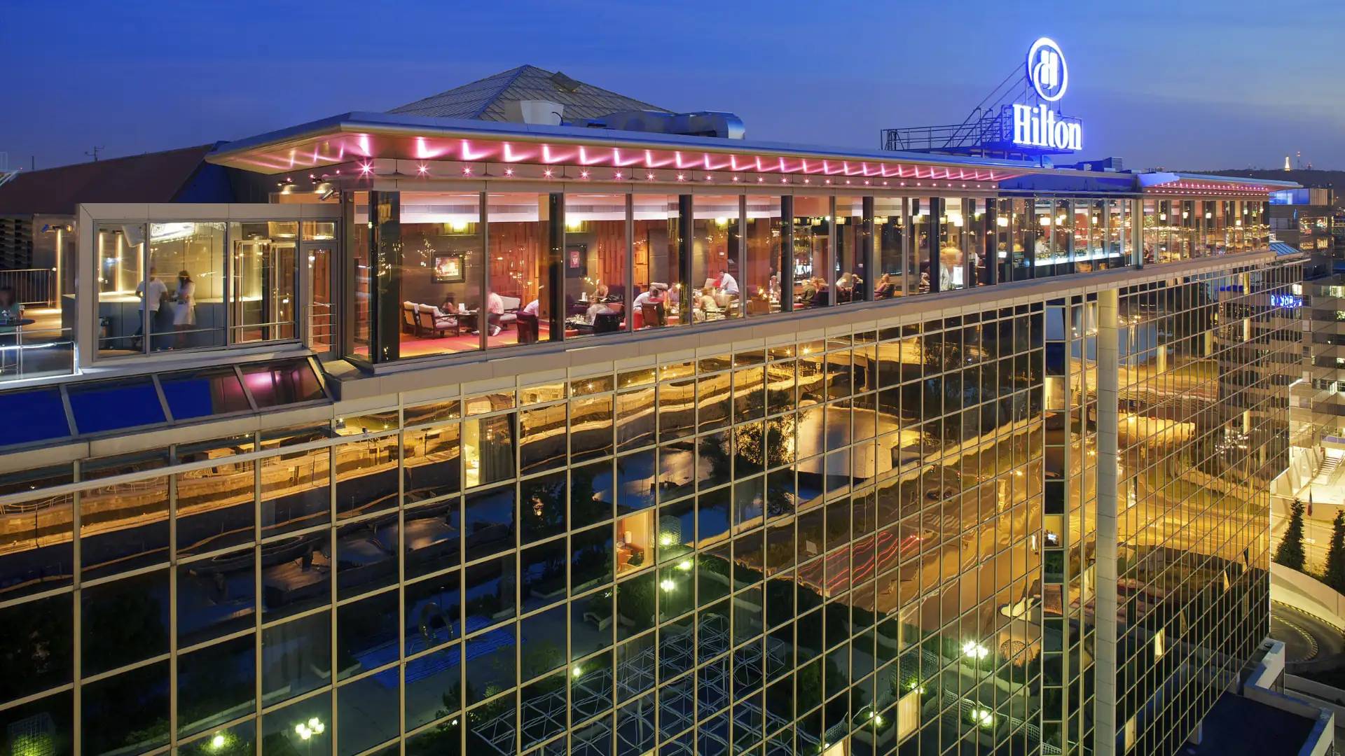 Hilton Prague Außenansicht im Kachelformat: Hilton Hotel in Prag bei Abenddämmerung mit beleuchtetem Logo und verglaster Fassade.