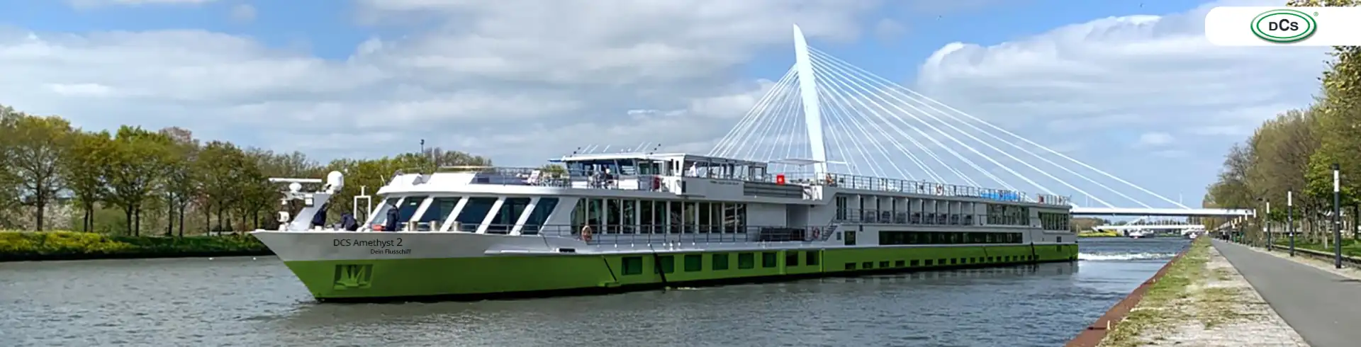 DCS Amethyst 2: Flusskreuzfahrtschiff mit grün-weißer Gestaltung auf einem Kanal vor einer modernen Hängebrücke und Baumreihe (Bannerformat)