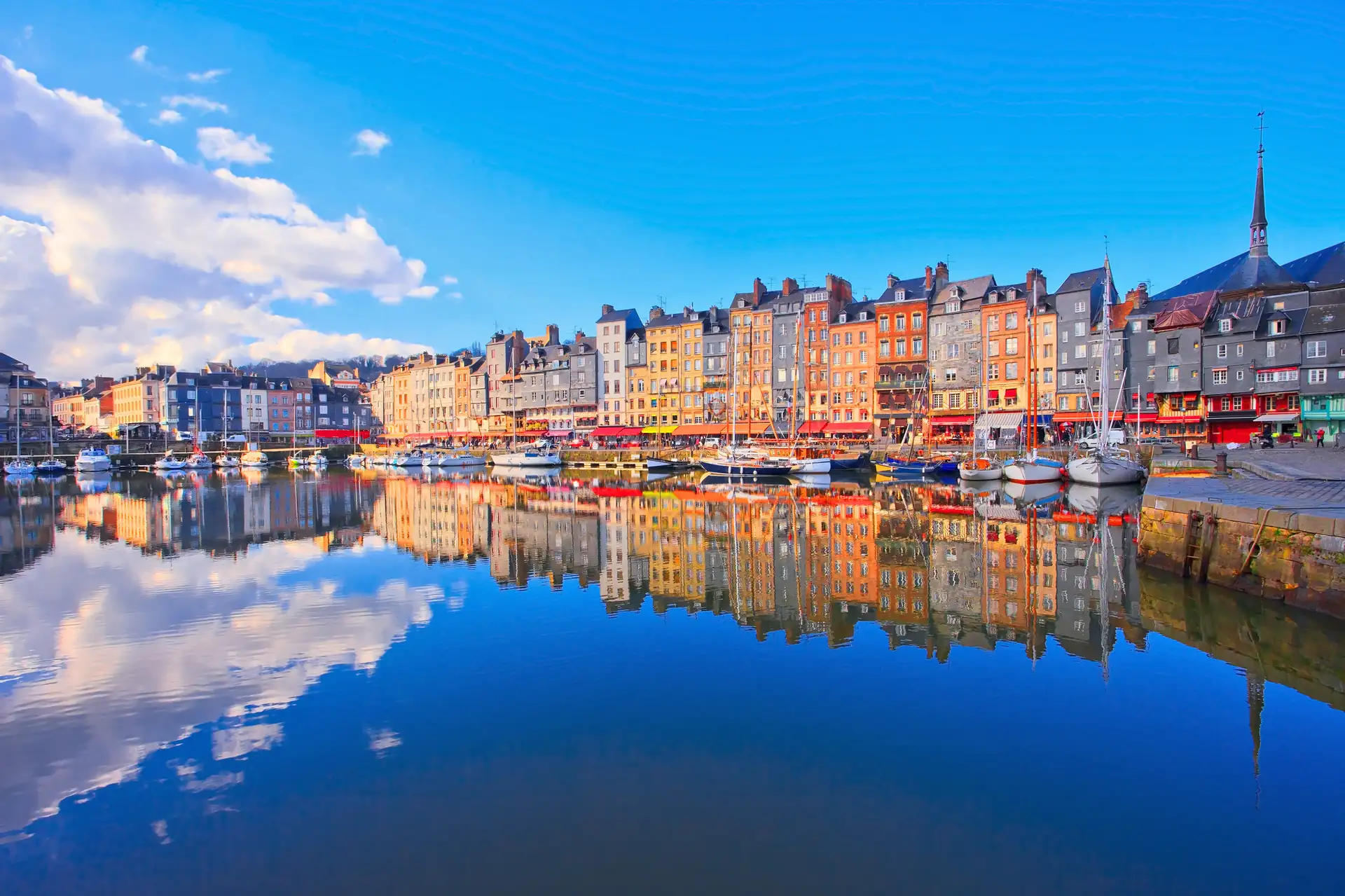 Honfleur: Spiegelung von bunten Häusern und Segelbooten an einem Hafen unter blauem Himmel mit Wolken.