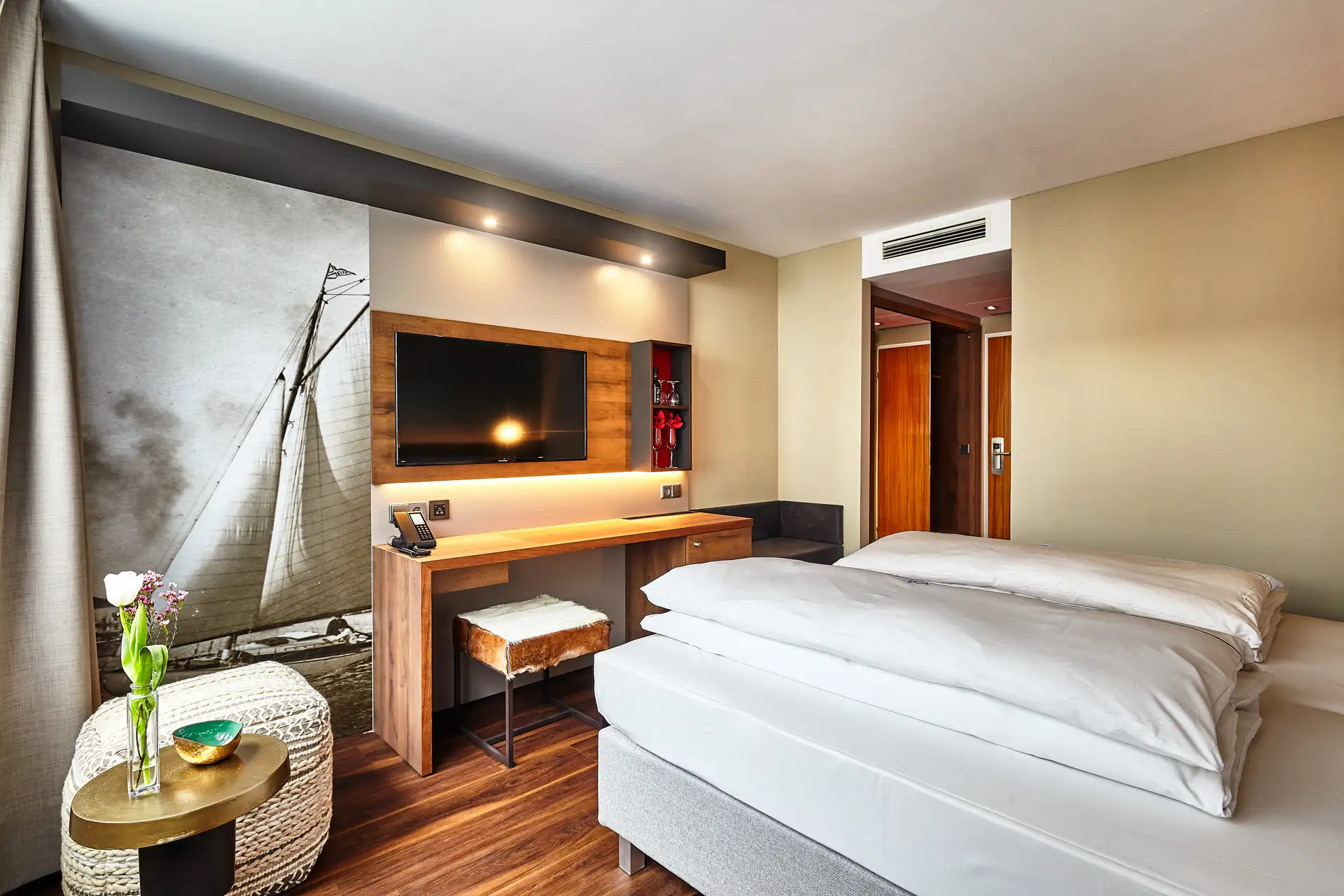Doppelzimmer im Hotel City Krone mit einem großen Bett, einem Schreibtisch mit Fernseher und einer dekorativen Wand mit einem Segelboot-Motiv. Blumen in einer Vase auf einem Tisch.