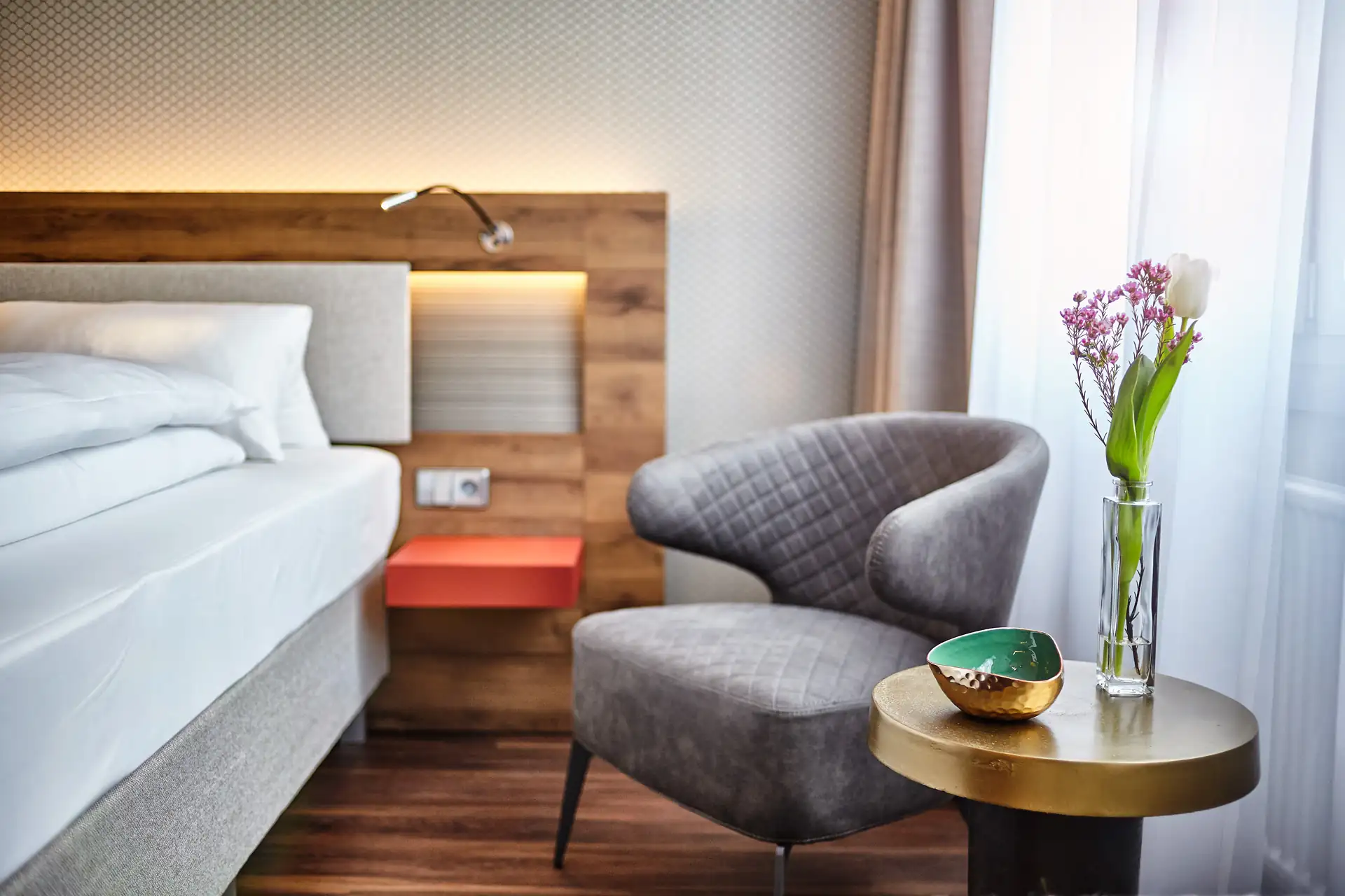 Doppelzimmer im Hotel City Krone mit einem Bett, einem grauen Sessel und einem kleinen Tisch. Auf dem Tisch steht eine Vase mit Blumen und eine Schale.